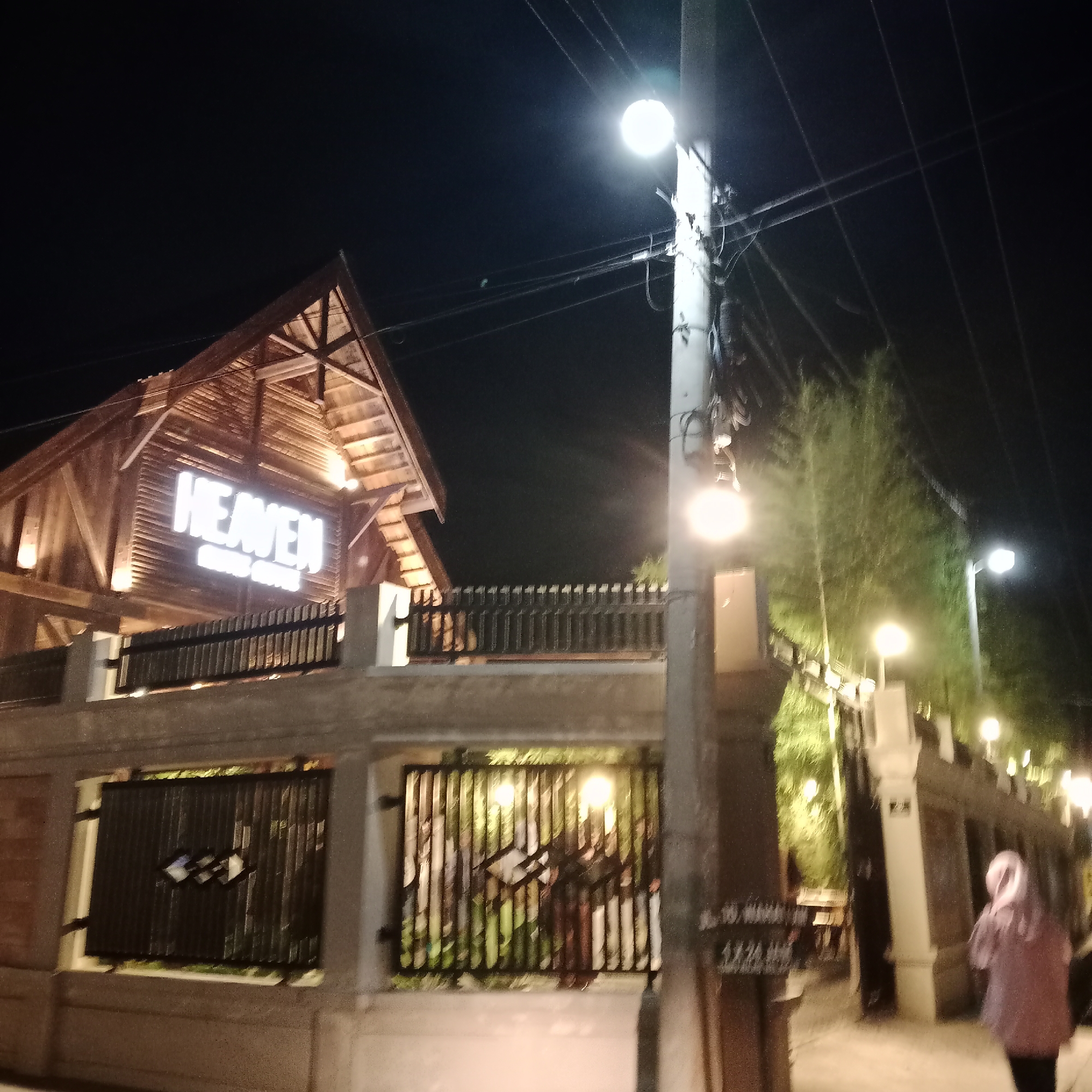 The image captures a nighttime scene in Bireuen Meunasah Dayah, Indonesia, with a focus on a wooden building and its illuminated signage. The building has a prominent "HEAVEN" sign in large, white, illuminated letters, with smaller text beneath it that is difficult to decipher. The structure itself appears to be made of wood, with a peaked roof and visible rafters. A fence made of dark metal bars encloses the area in front of the building, and further back, more fencing and some greenery are visible, also illuminated by lights.
A street lamp with a bright light is visible on the right side of the image, attached to a utility pole with numerous wires. Another street lamp is visible further down the path. The lighting suggests it is nighttime. In the lower right of the frame, a person wearing a pink hijab and a pinkish-purple jacket is walking away from the camera, their back to the viewer. Their face is not visible, and their posture does not convey any specific emotion. The overall mood is quiet and somewhat atmospheric due to the nighttime setting and the lighting. There are some indistinct signs on a pillar to the right of the person, but the text is illegible. The weather appears clear, with no visible rain or fog.