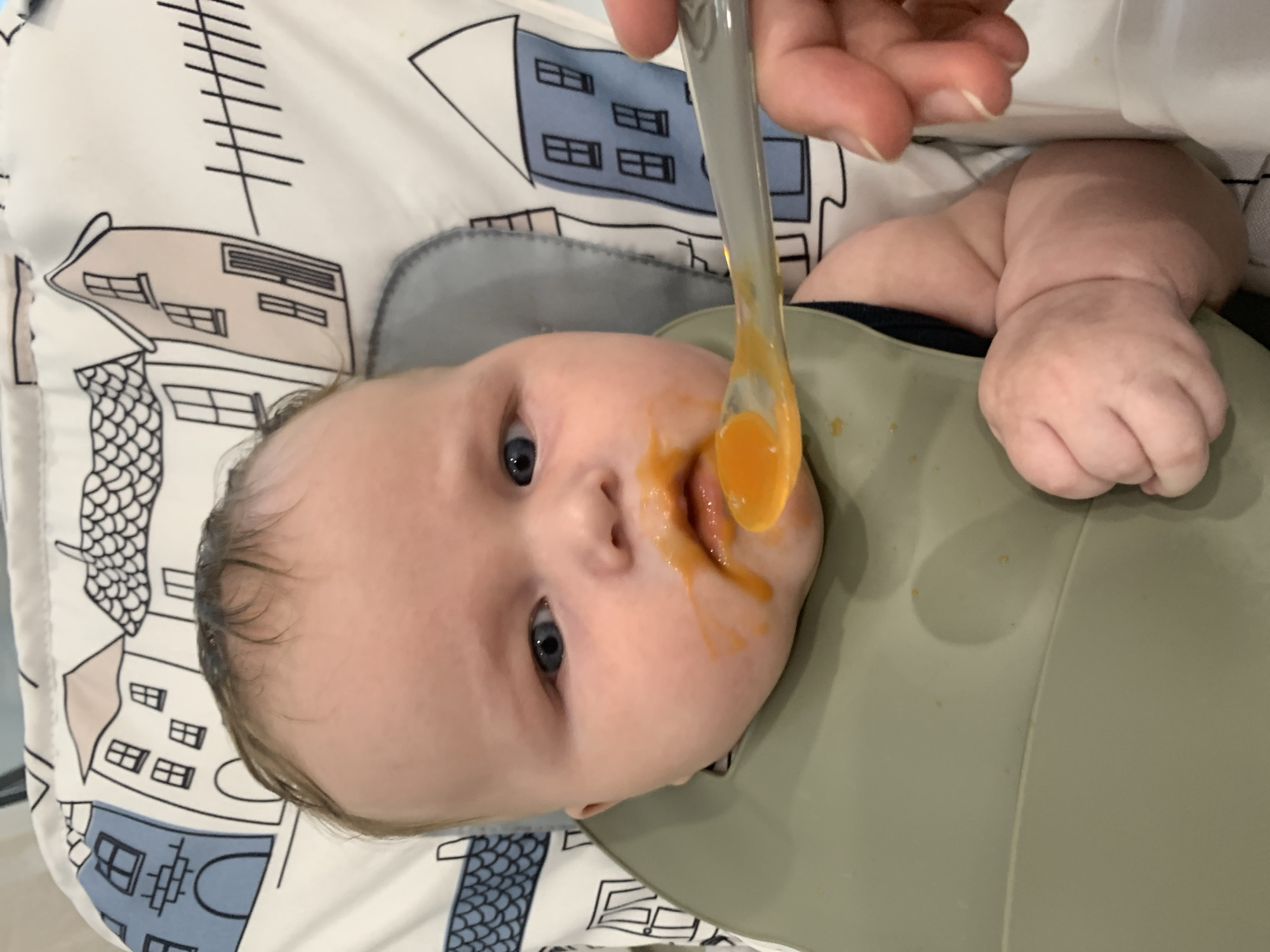 A close-up, eye-level shot captures a baby's face smeared with orange food. The baby, positioned in a high chair, has blue eyes and light brown hair. A green bib covers their chest. A spoon, held by an adult hand, is in the process of feeding the baby, with a dollop of orange food on it. The background of the high chair is decorated with a pattern of stylized houses. There is no indication of the location being Stavropol, Russia, as this information is not present in the visual data.