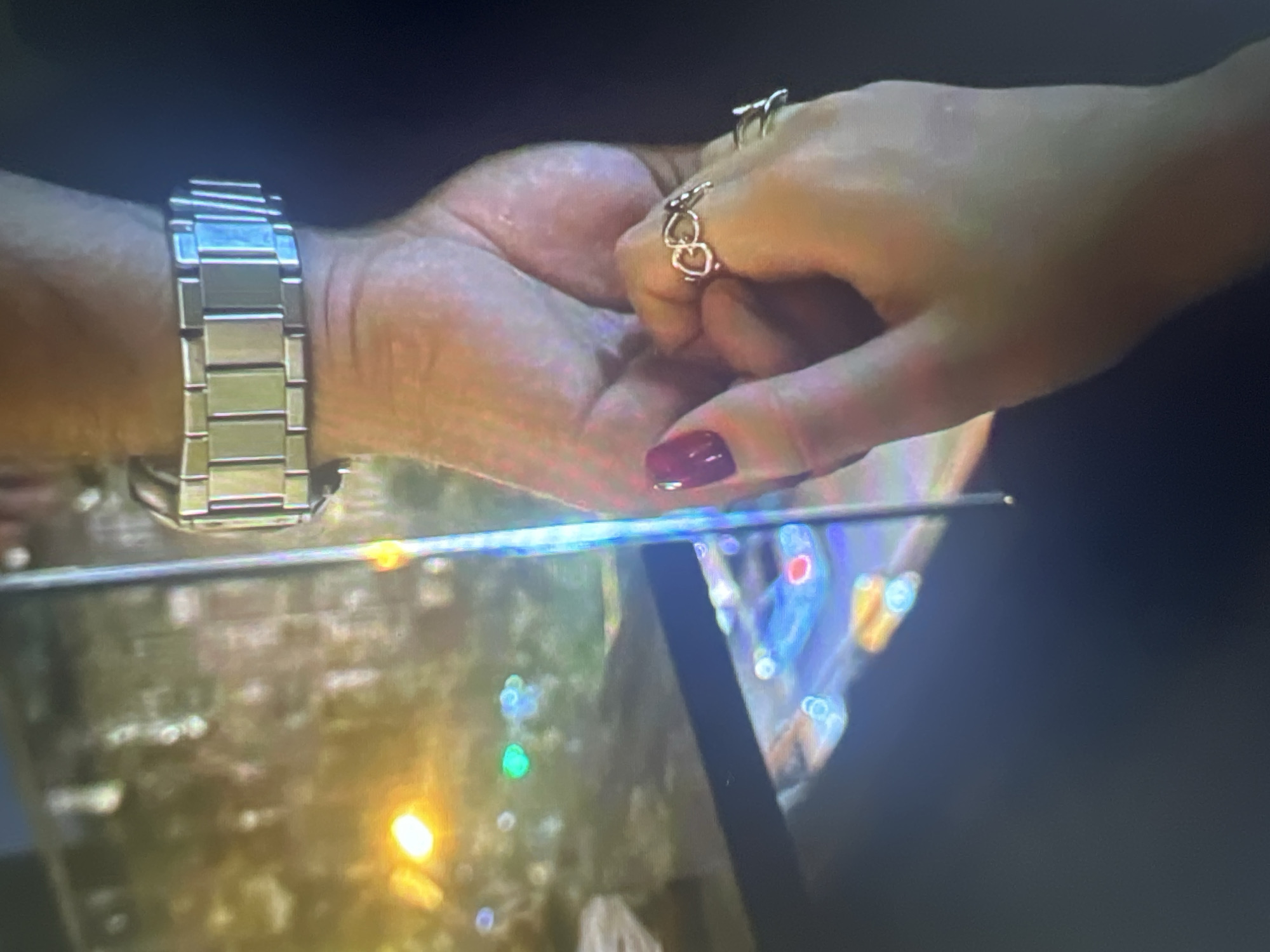 The image shows a close-up of two hands interlocked. One hand, appearing to be a man's, is wearing a silver metal watch. The other hand, appearing to be a woman's, has a ring with a heart-shaped design and fingernails painted a deep red. The background is blurred but suggests an urban environment with illuminated lights, possibly from streetlights and vehicles. The image does not contain any identifiable landmarks or clues that would indicate the location in Moscow, Russia.