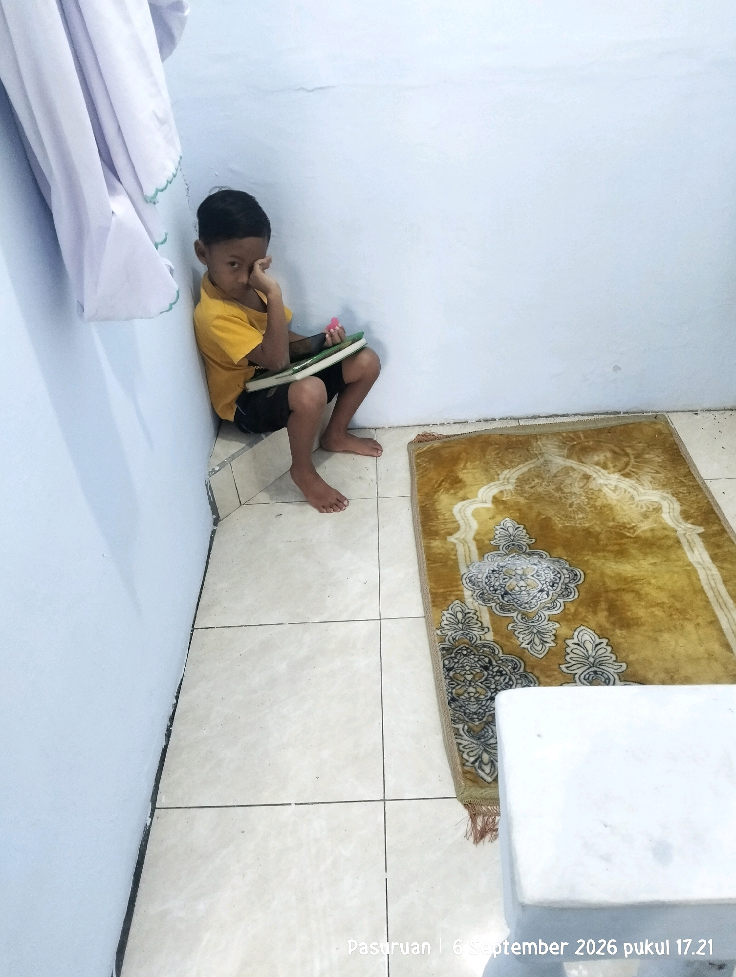 In the city of Warung Dowo, Indonesia, a young boy sits in a corner, leaning against a light blue wall. He is wearing a yellow t-shirt and black shorts. His left hand is touching his face, and he holds a book or tablet in his right arm. A prayer mat with a gold and white patterned design lies on the beige tiled floor to his right. A white object, possibly a small table or stool, is partially visible in the lower right corner of the image. The scene is indoors, with a white curtain hanging on the wall to the boy's left. The time stamp on the image indicates it was taken on September 6, 2026, at 17:21.
