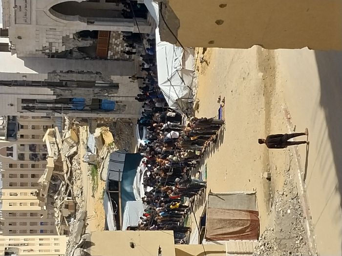 The image displays an outdoor urban scene in Khan Yunis, Palestinian Territory, characterized by significant destruction and a large gathering of people. In the mid-ground, a substantial group of individuals, predominantly adult males, is arranged in multiple rows on an unpaved dirt surface, performing a collective prayer; some utilize individual prayer mats, with shoes placed nearby. A lone individual in dark clothing stands with his back to the camera in the immediate foreground, observing the scene. To the right, a prominent white mosque structure with an arched entrance and a partial minaret shows extensive structural damage, including large sections of its facade missing, exposing interior elements, with blue tarpaulins visible within damaged openings. In the background, vast piles of rubble and debris from demolished buildings are evident, with several multi-story residential buildings, some visibly damaged, standing further beyond. White tarpaulin shelters are visible in the mid-ground, and several white vehicles are partially obscured amongst the crowd. The ground consists of unpaved dirt and scattered debris. The scene occurs under daylight with a clear sky.