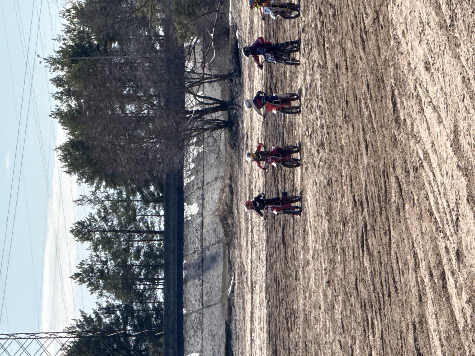 The image captures a motocross race in Chaikovsky, Russia, on a dirt track. Several riders on dirt bikes are seen in varying positions, suggesting active racing. The track surface is textured, displaying signs of recent activity from tires. A line of trees and utility poles runs parallel to the track on one side, while the backdrop includes a clear sky. The composition suggests a natural landscape setting with man-made elements for the event.