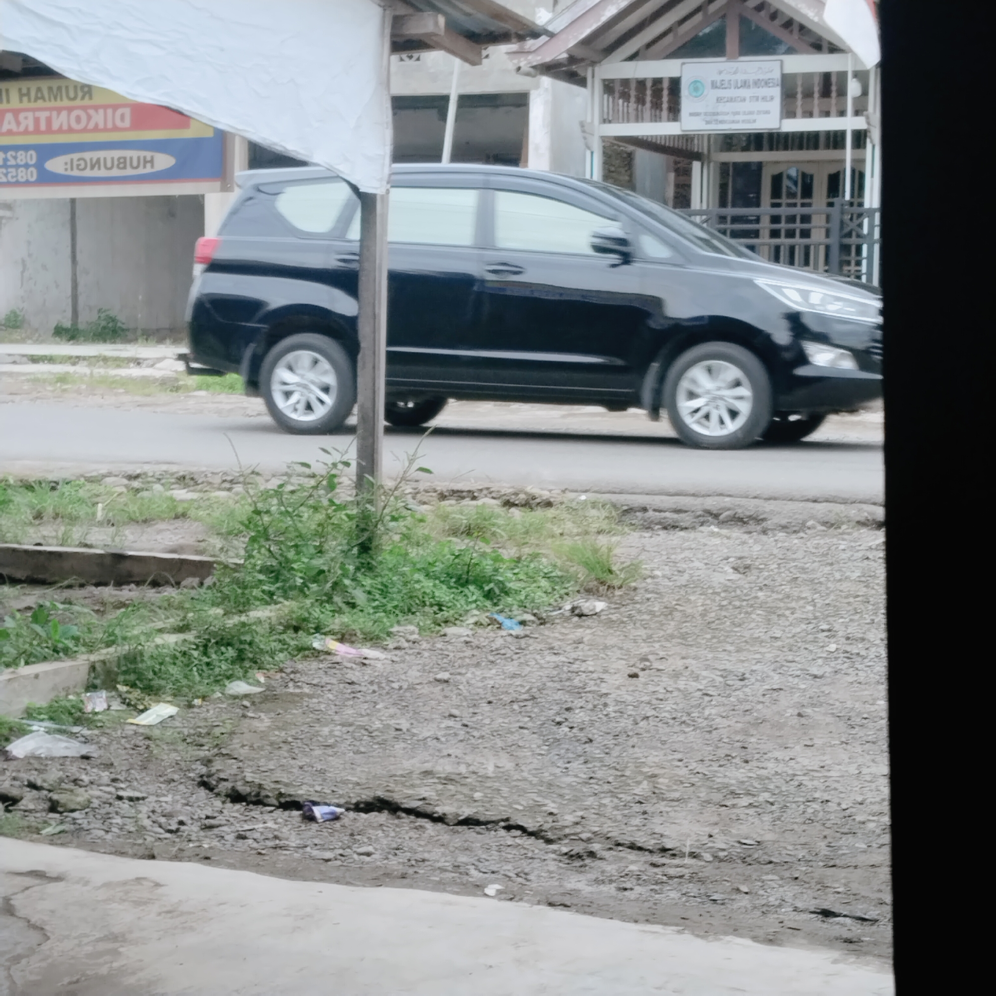 The image captures a street scene in Talun Kenas, Indonesia. A black SUV is parked on the side of a paved road. In the background, a building with a tiled roof and wooden supports is visible, with a sign that reads "MAJELIS ULAMA INDONESIA" and "KECAMATAN TIMUR ILIR." A white tarp is draped over some wooden structures in the foreground. The ground alongside the road is a mixture of dirt, gravel, and patchy grass, with some scattered debris, including plastic wrappers. The overall environment appears to be somewhat unkempt.