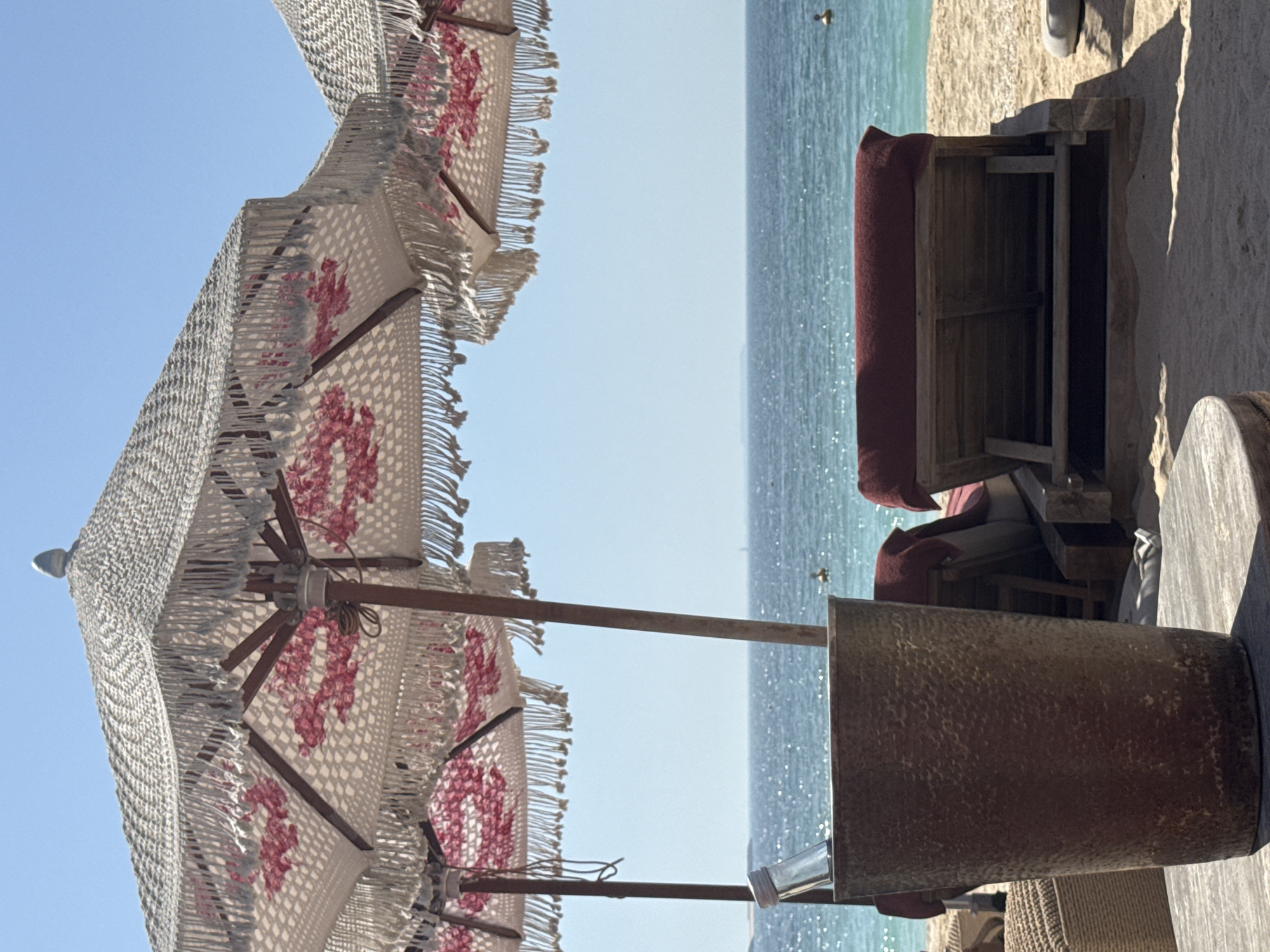 The image shows an outdoor beach scene, likely at a resort or private beach. In the foreground, there is a textured bucket or container on a table, along with part of a beach umbrella with decorative fringe and a pattern that may include pink flowers. Beyond these, there are beach lounge chairs or daybeds with dark red cushions, positioned on sand. In the background, the sea merges with a clear, light blue sky. The water is calm with minor glimmers. The overall impression is of a serene, sunny day at a luxurious beach location. Given the description, it is located in Dubai, United Arab Emirates.