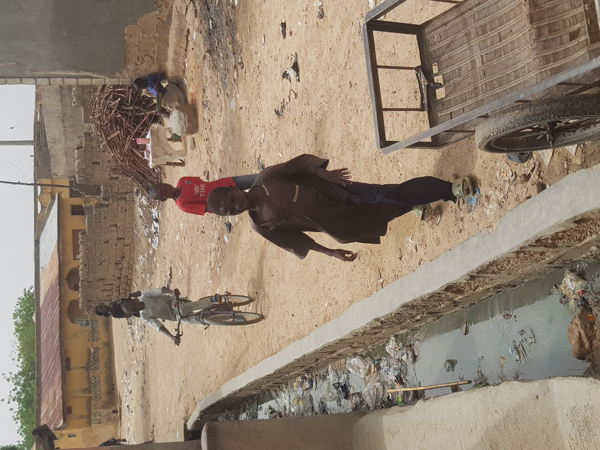 Location: Dutsinma B, Nigeria.
Scene: An unpaved street with sandy terrain.
People: A man in a brown garment stands next to a handcart, while another man in a red shirt is visible near a pile of sticks and debris. A third man is seen riding a bicycle further down the street.
Objects: A handcart with a wooden slat bed and a single tire is positioned to the right. Sticks and debris are collected near the first man in red. A drainage ditch filled with trash and stagnant water runs along the side of the street.
Actions: The man in brown appears to be observing the scene. The man in red is situated near the pile of sticks. The third man is riding his bike down the street.
Environment: The setting appears to be a residential area with buildings visible on either side of the street. The overall condition of the area appears unkempt, with trash and debris scattered throughout the scene.