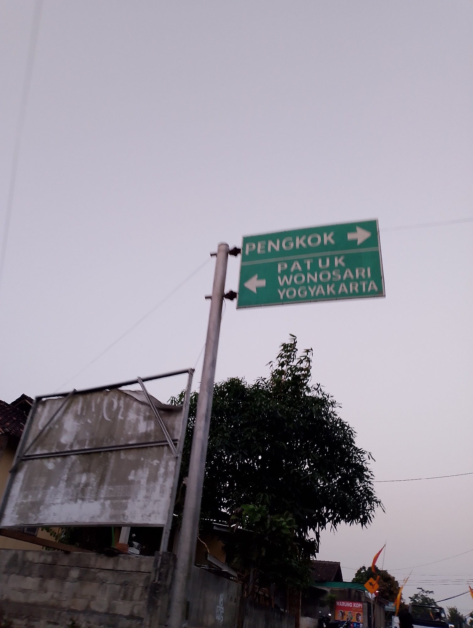 A traffic sign with two green panels is mounted on a pole in Patuk, Indonesia. The upper panel reads "PENGKOK" with an arrow pointing to the right. The lower panel reads "PATUK WONOSARI YOGYAKARTA" with an arrow pointing to the left. To the left of the sign, a large, dilapidated billboard is visible, supported by a metal frame. In the background, a dense tree obscures a portion of the sky, and further to the right, smaller trees and flags are visible. A brick wall runs along the bottom of the image. The sky is overcast.