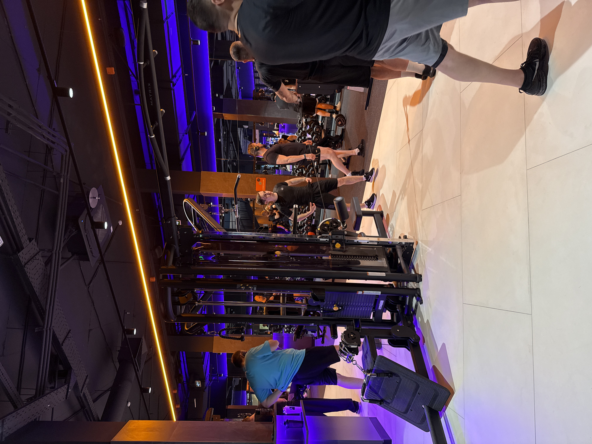 The image captures the interior of a gym. People are using exercise equipment. In the background are rows of dumbbells. The gym has a black ceiling with visible industrial elements. There is a bright yellow light. Blue light illuminates the walls. The image was likely taken indoors. It is difficult to determine if the location is in Krasnoyarsk, Russia from the image.