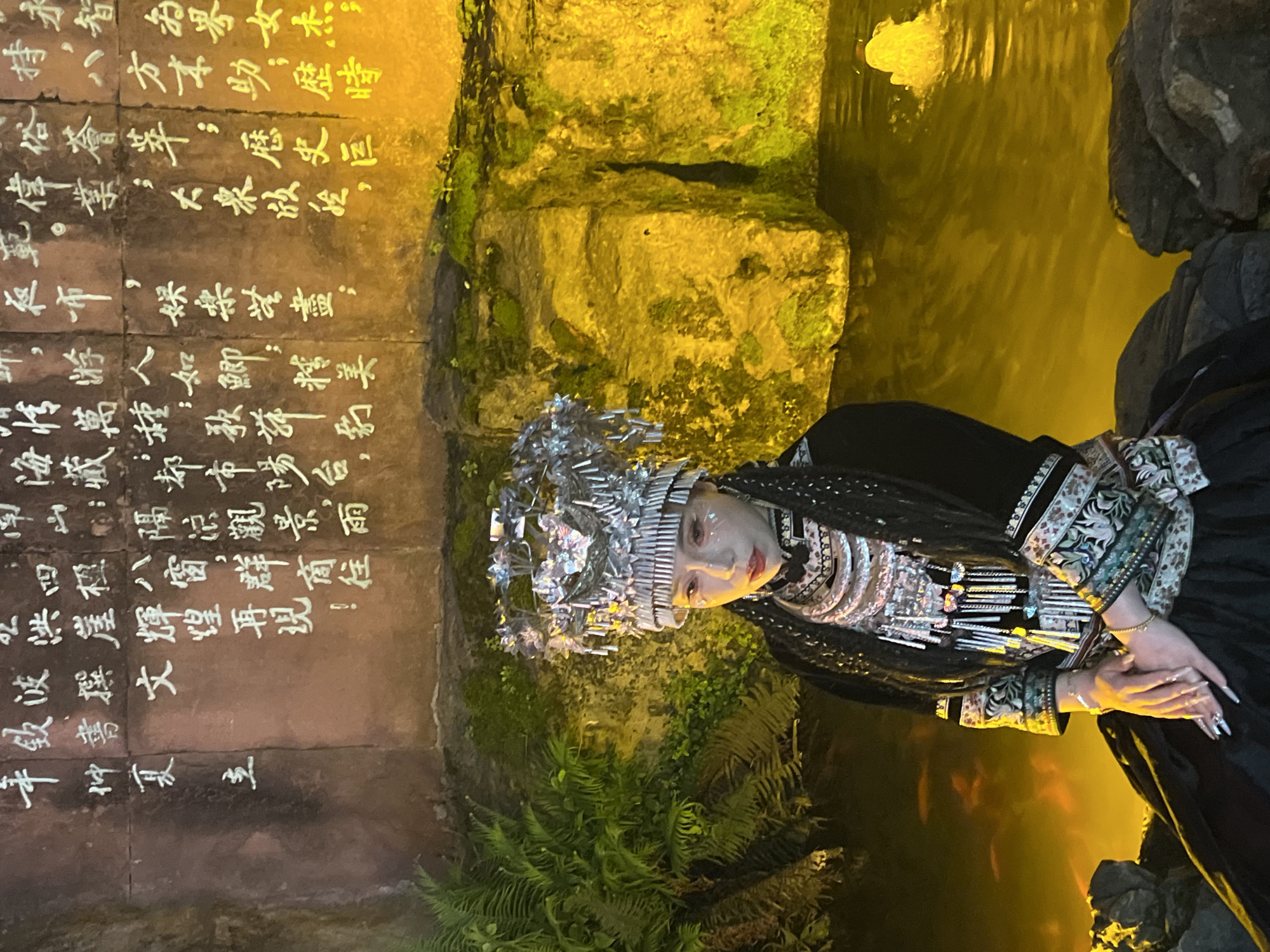 The image features a woman adorned in traditional Miao attire, including an elaborate silver headdress and ornate clothing, posed against a backdrop of a stone wall inscribed with Chinese calligraphy. The setting appears to be a grotto or alcove illuminated with warm, golden light. The environment includes natural elements like water and foliage, suggesting an outdoor or landscaped location. The presence of the calligraphy and traditional clothing point to a cultural or historical setting. The location is likely in the Yuzhong District of China.