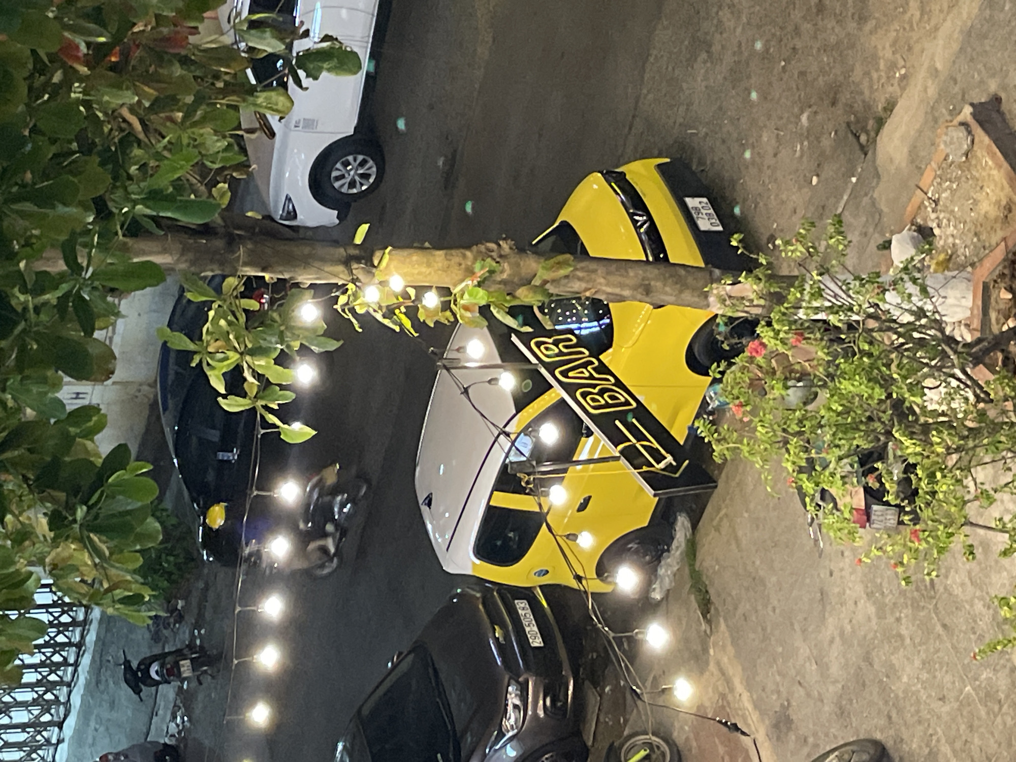 From an elevated viewpoint, the image captures a nighttime street scene in Bắc Nha Trang, Vietnam. The perspective looks down onto a paved road lined with parked vehicles and vegetation.
A prominent tree with a thick, textured trunk stands roughly in the center, acting as a focal point. Decorative string lights with warm, glowing bulbs are wrapped around its trunk and branches, illuminating the immediate surroundings. Lush green foliage, likely from the tree and other plants, frames parts of the shot.
Several vehicles are parked along the street. To the left (when viewing upright), a white car is partially visible, with its front wheel and a section of its bumper peering from behind foliage. Further right, a distinctive yellow and white vehicle, possibly a small delivery van or taxi, is parked. The side of this vehicle displays the bold text "BAR" in black against its yellow body. Its rear license plate, "79A 038.02", is clearly legible, indicating its registration in Khánh Hòa province. Behind this vehicle, a small bush with a few red blossoms grows on the ground.
In the foreground, a dark grey SUV is parked. Its front license plate, "29D 505.83", is also clearly visible, identifying it as another Vietnamese vehicle. Several motorbikes are scattered throughout the scene, one partly obscured by the SUV, another near the white car, and a third further down the street.
The overall lighting suggests it is evening or night, with the string lights providing localized illumination and other artificial lights glowing softly in the background. The scene depicts a typical urban street, relatively calm, with cars and motorbikes parked along the curb amidst greenery.