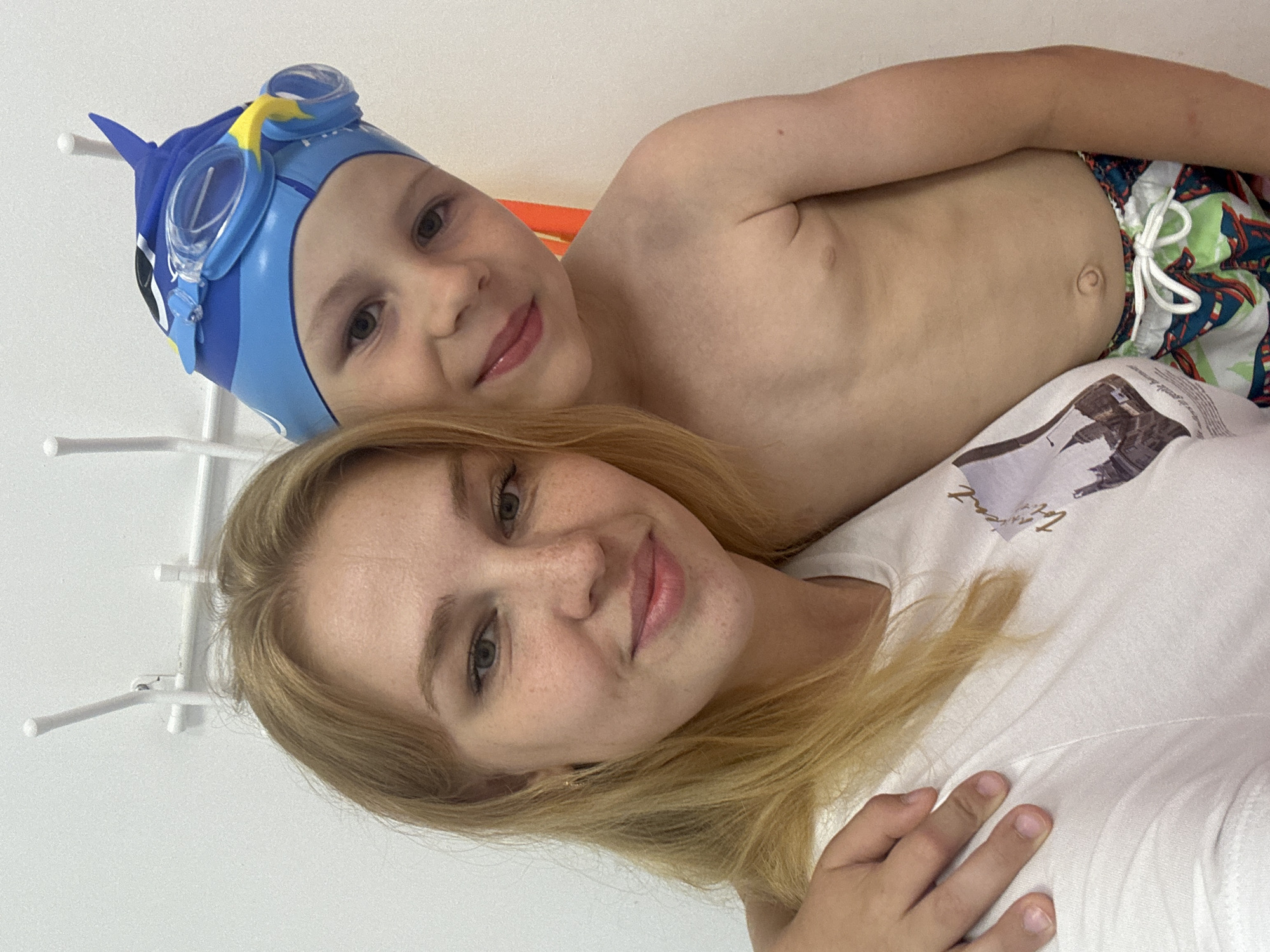 The image shows a close-up, vertically oriented selfie of two people, likely a mother and her young son, in what appears to be an indoor setting, possibly a changing room or a poolside area in Rusanovo, Russia. The woman in the foreground has blonde hair, fair skin with freckles, and is wearing a white t-shirt. She is smiling, with her head tilted slightly. A young boy, wearing a blue swimming cap with goggles and colorful swimming trunks, is positioned behind her and slightly to the side. He is also looking at the camera and appears to be smiling. The background is a plain white wall with some white horizontal bars. The lighting suggests it is daytime. The overall mood is happy and casual, indicative of a day spent swimming or at a pool. There is no visible text in the image.
