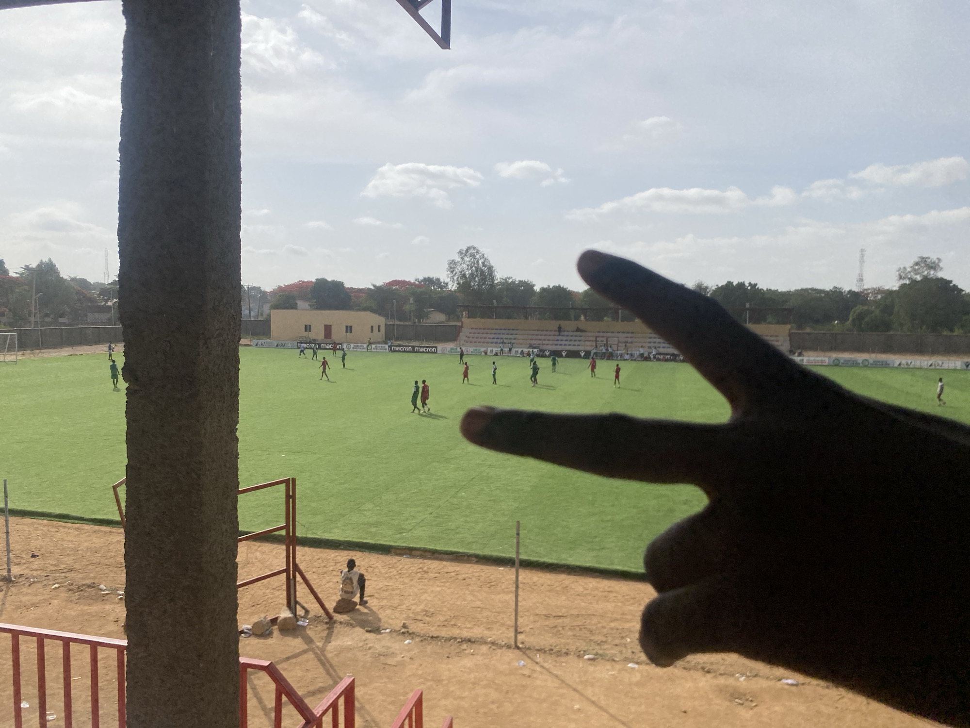 The image shows a football match in progress at a stadium in Tudun Wada, Nigeria. The stadium has a green artificial turf field with players in red and green jerseys. The spectator's view is partially obstructed by a concrete pillar and their hand in the foreground. There are buildings and trees visible beyond the stadium. A man sits on the ground below the stadium.