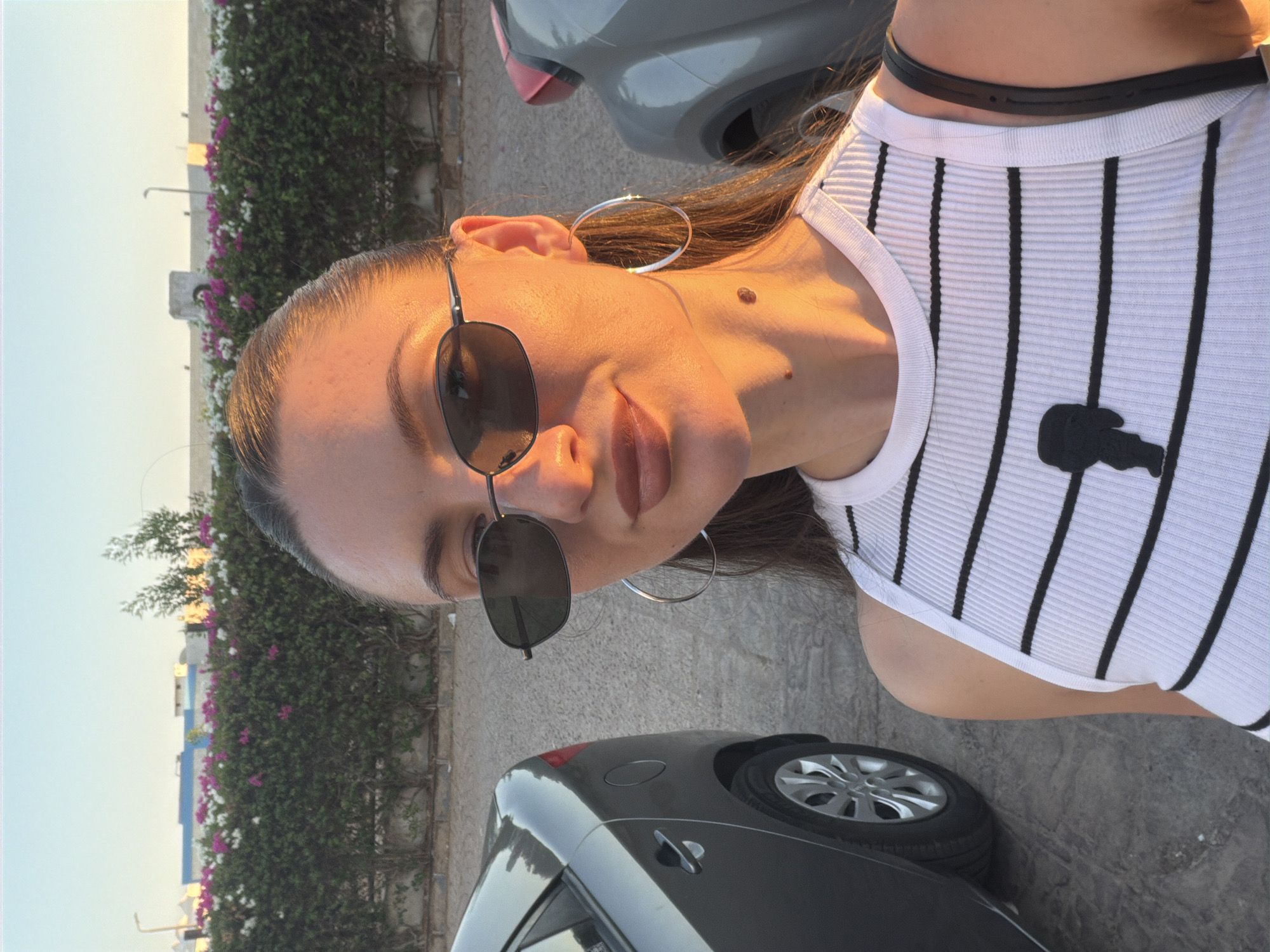 A woman is taking a selfie outdoors in Sharm El Sheikh, Egypt. She is standing in a parking lot, with cars on either side of her. Behind her is a hedge with pink and white flowers. The sun is setting, casting a warm glow on her face. She is wearing sunglasses, a white and black striped tank top, and large hoop earrings. The background shows buildings and a clear sky.