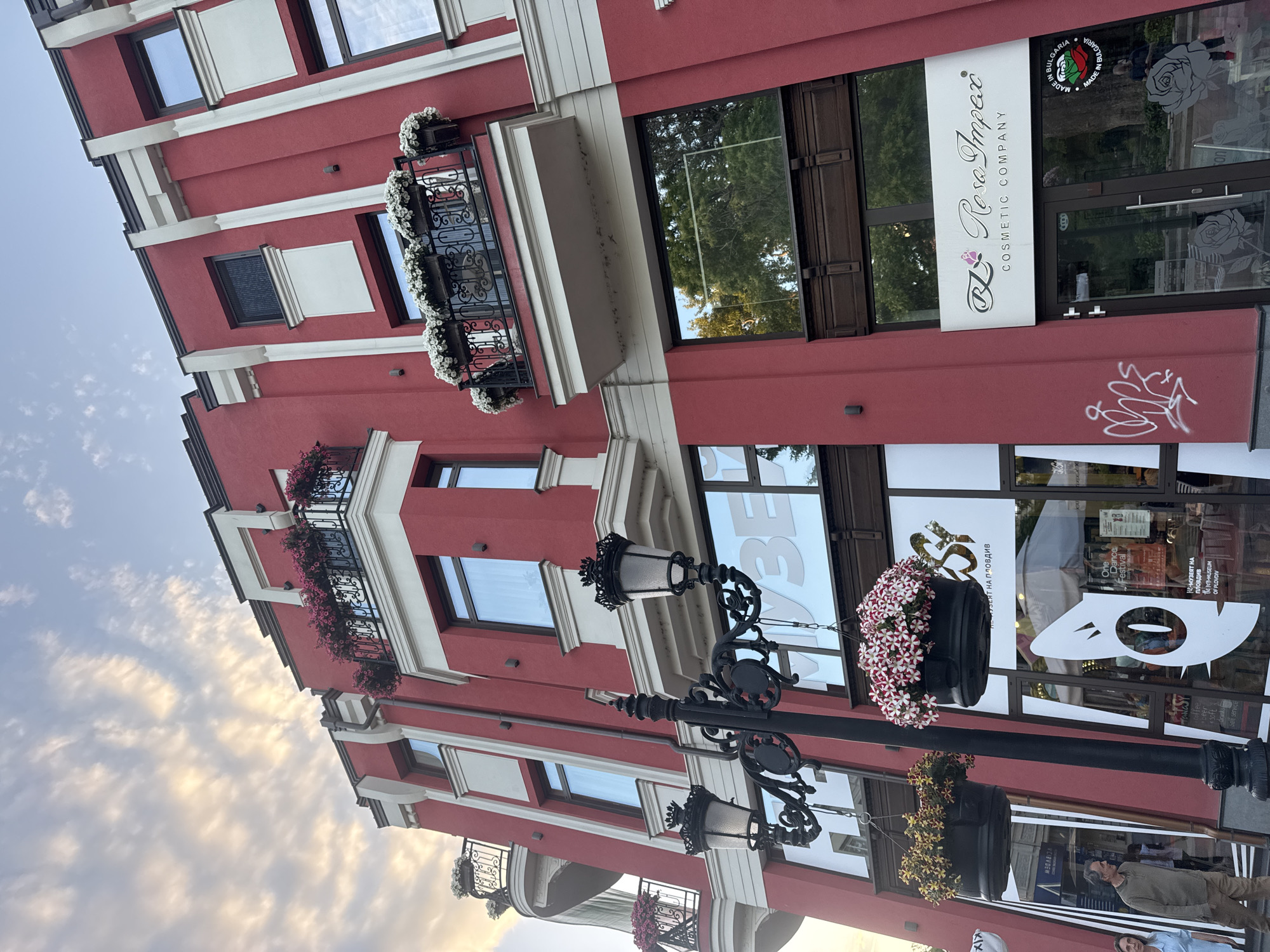 The image shows a street scene in Plovdiv, Bulgaria. A three-story red building with white trim and wrought-iron balconies adorned with flowers stands prominently. The ground floor hosts a shop or business named "Rosa Impex Cosmetic Company", with signage visible on the storefront. Additional signage indicates the location of the "N-Museum of Plovdiv" and the "One Dance Festival". Street lamps with hanging flower baskets are placed in front of the building. A man is visible on the street, and the sky is partially visible in the background.
