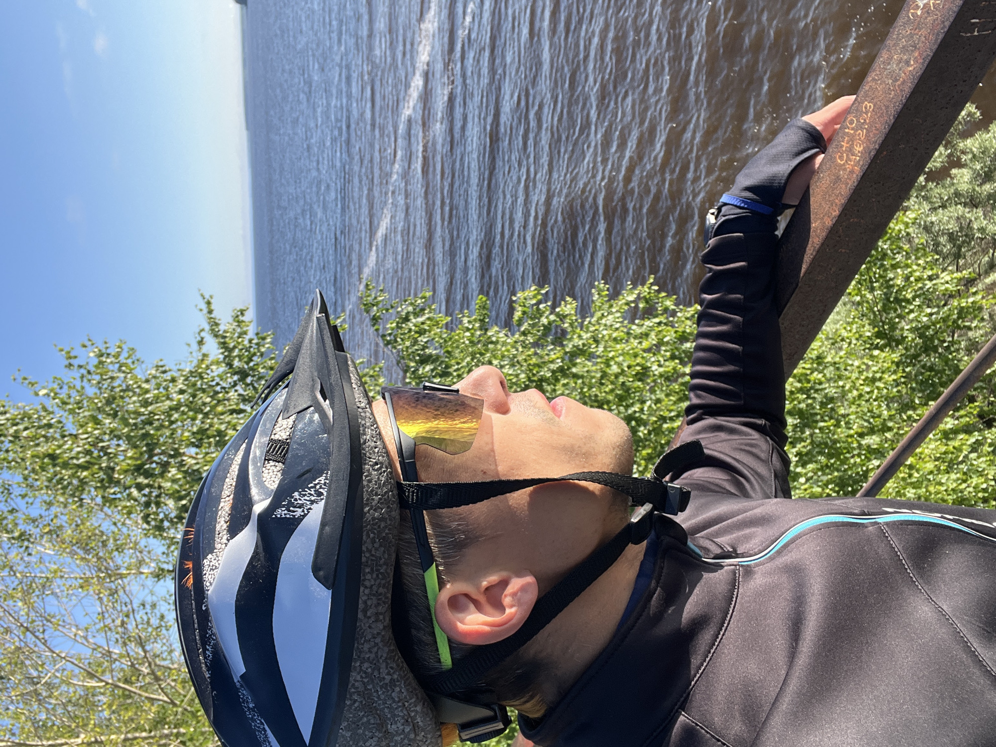A man wearing a black cycling jacket, helmet, and reflective sunglasses is leaning on a rusty metal railing, looking out at a wide body of water. The water appears to be a lake or river, with ripples on its surface. Trees with green leaves are visible in the foreground and background. The sky is clear and blue, with a hint of haze on the horizon. The scene is lit by natural sunlight, casting shadows on the man and the railing. The man's right hand rests on the railing, revealing a blue band on his wrist. The railing has some faint markings on it. This scene is likely located along the Kama River in Chaikovsky, Russia, given the characteristic landscape and vegetation.