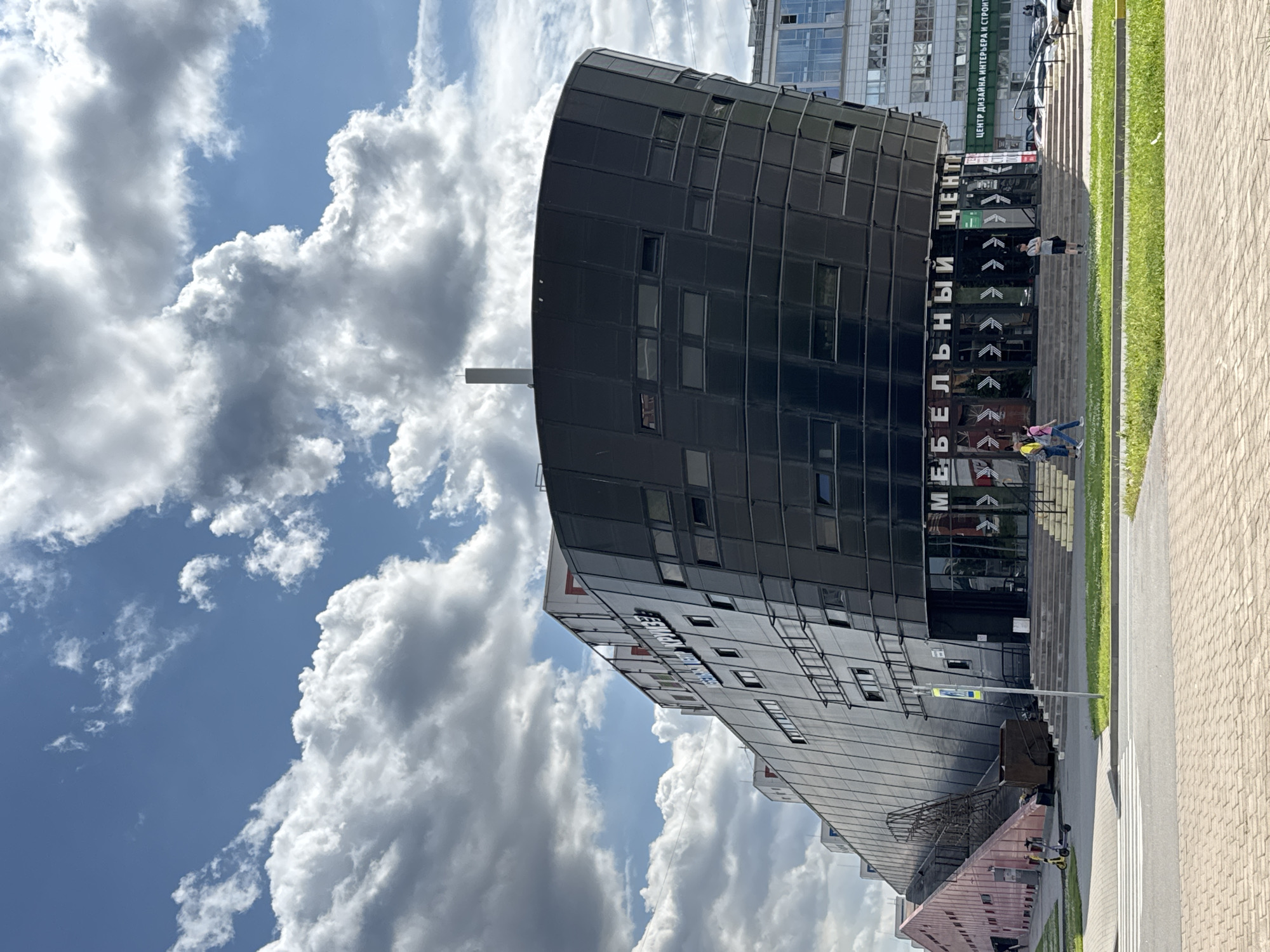 The image shows a modern, dark-colored, curved building with many windows under a bright blue sky with fluffy white clouds. The building has an entrance with a set of stairs leading up to it. A few people are walking up the stairs and near the entrance. The building has text on it that reads "МЕБЕЛЬНЫЙ ЦЕНТР," which translates to "Furniture Center." Another sign visible above the entrance reads "ЦЕНТР ДИЗАЙНА ИНТЕРЬЕРА И СТРОИТЕЛЬСТВА," which translates to "Center for Interior Design and Construction." To the right of the building, there is a sidewalk with a brick pattern and a grassy area. In the foreground, there is a pedestrian crossing. A dumpster and some small vehicles are visible on the pavement near the building's lower section. The image appears to be taken during the day, with bright sunlight casting shadows, indicating a sunny weather. The setting appears to be an urban area in Saint Petersburg, Russia, given the text on the building.
