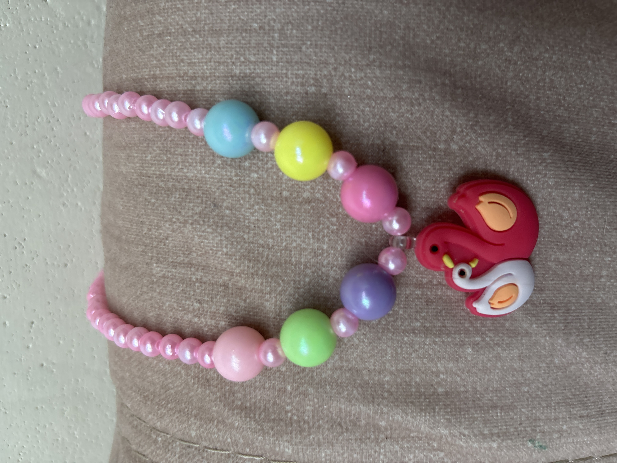 A pink beaded necklace with a swan pendant is displayed on a textured, light brown fabric surface. The necklace features a series of larger, round beads in various pastel colors including pink, green, purple, blue, and yellow, interspersed with smaller pink pearlescent beads. At the bottom of the necklace is a bright pink pendant shaped like two swans, one larger and one smaller, with white detailing on the smaller swan's neck and orange accents. The background is a textured, light-colored wall. The image does not contain any information or clues regarding the city of Imeni Morozova, Russia.