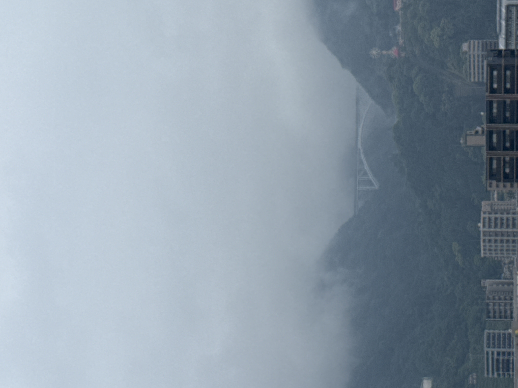 The image depicts a cityscape of Beppu, Japan, shrouded in fog and captured from an elevated perspective. The predominant feature is the dense fog obscuring the landscape, yet details such as the suspension bridge and the verdant mountainside are partially visible through the mist. The right side of the image showcases the urban development of Beppu with a cluster of high-rise buildings, constructed in a box-like architectural design, suggesting residential or commercial structures. The buildings are positioned at the edge of the mountain range, indicating an integration of urban and natural environments. The absence of distinct human figures implies that the scene emphasizes the environment over human presence.
