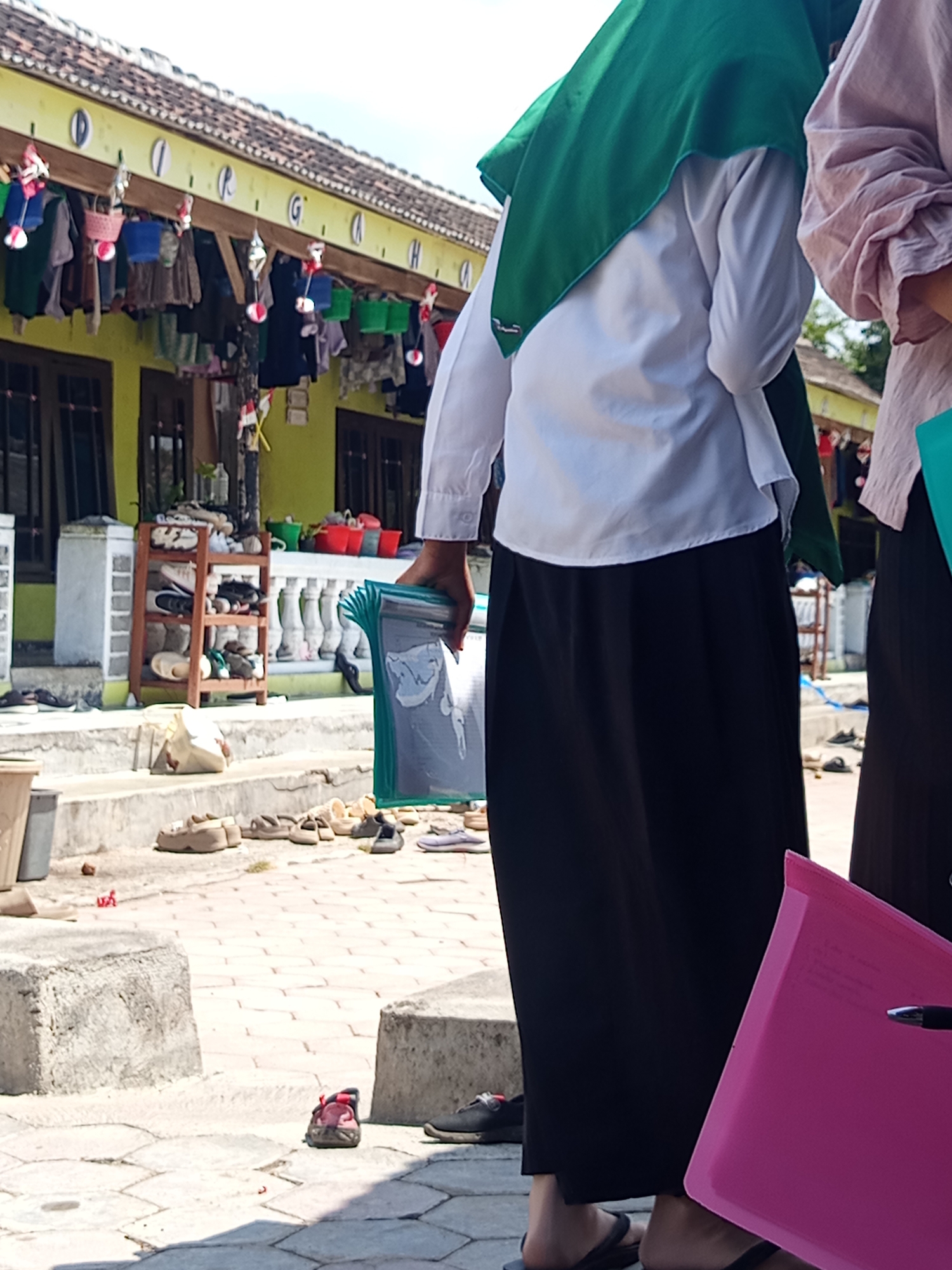 In Lumutan, Indonesia, two young women, presumably students due to their uniforms, stand on a paved outdoor area. The woman on the left wears a white long-sleeved shirt and a dark skirt, her face obscured by a green hijab. She holds a stack of green plastic folders. The woman on the right, partially visible, wears a pink, loosely fitting garment, also with her head covered.
The background suggests a modest building with a yellow facade, a tiled roof, and a row of windows. A wooden shoe rack filled with various footwear is positioned against the building. Clothes hang from a line under the eaves of the roof, adorned with decorative elements. The ground is paved with hexagonal tiles and large concrete blocks. Scattered footwear, including sandals and flip-flops, are visible on the ground and near the building. The overall scene depicts an everyday outdoor setting likely associated with a school or community area in Lumutan.