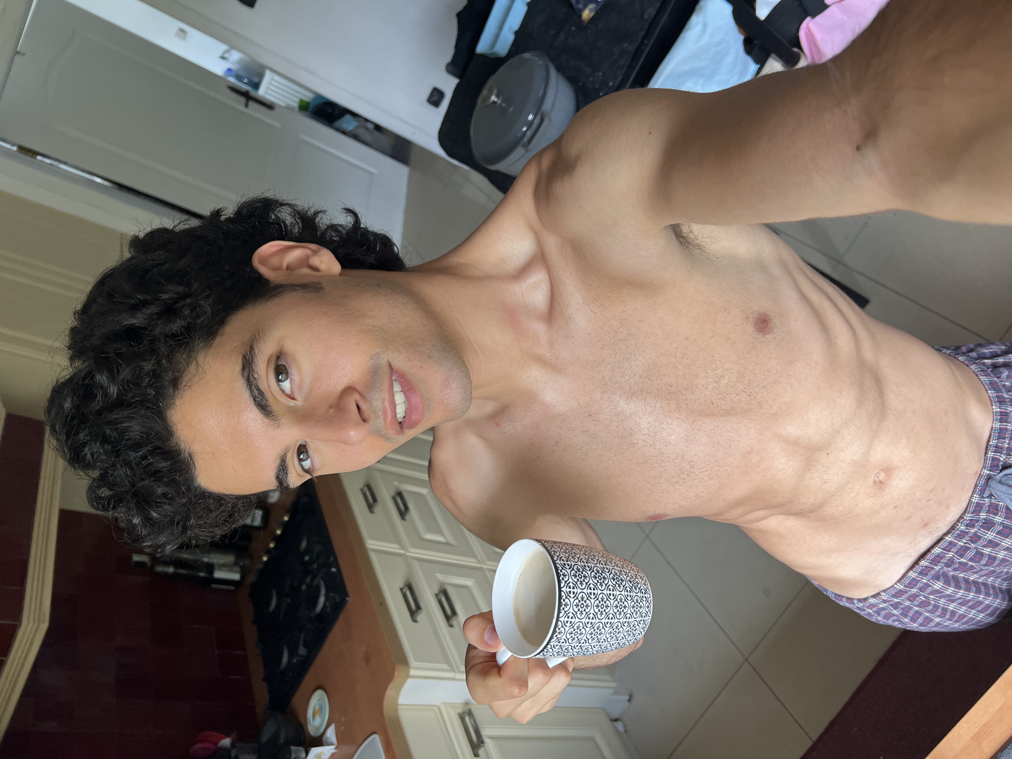 A shirtless man with curly dark hair and a patterned mug of coffee stands in a kitchen. The man is holding the mug in his left hand and smiling at the camera. He is wearing plaid boxer shorts. The kitchen has white cabinets, a black stovetop, and a brown countertop. The image is taken from a slightly high angle.