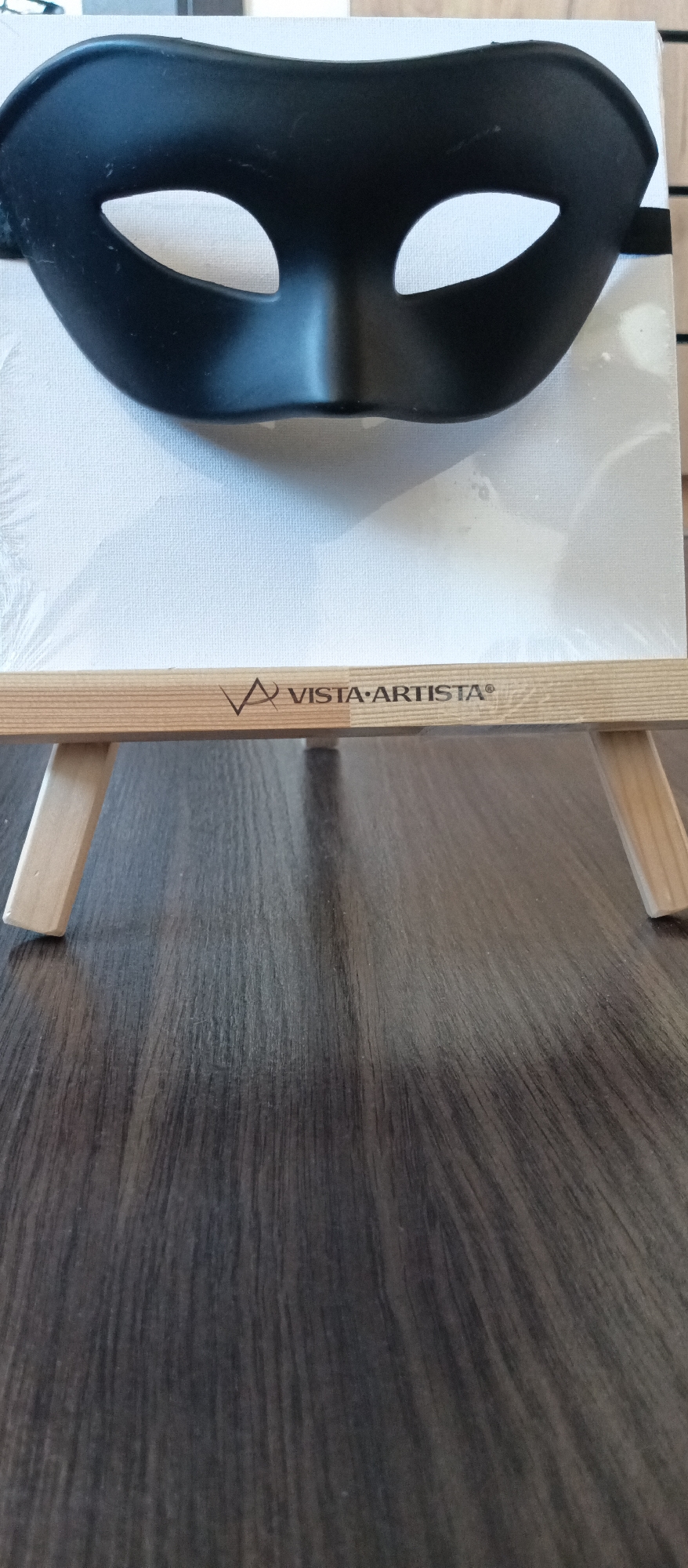 A black masquerade mask rests on a small, wrapped canvas set on a wooden easel. The easel's horizontal bar bears the logo "VISTA ARTISTA". The mask is positioned near the top of the canvas. The background consists of a dark, textured surface, likely a wood table. The location is Unknown, Unknown.