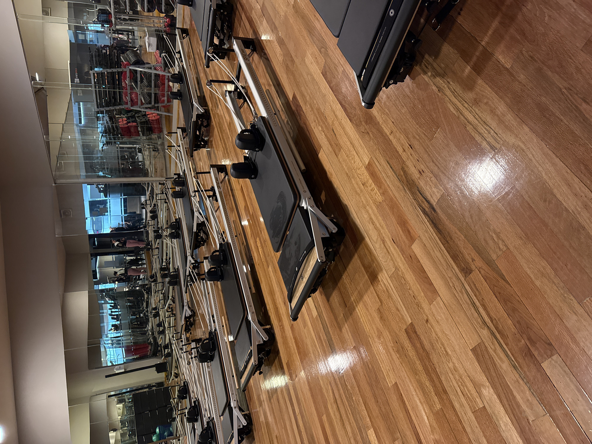 The image depicts a pilates studio in an unidentified location in Puebla, Mexico, evidenced by the interior design of the fitness gym. Rows of pilates reformer machines are arranged parallel to each other on a glossy hardwood floor. A mirrored wall stretches along one side of the studio, reflecting the machines and other equipment in the room. People are visible in the mirror's reflection, seemingly using the exercise equipment in the adjacent section of the room. The machines feature adjustable foot straps and sliding carriage beds.
