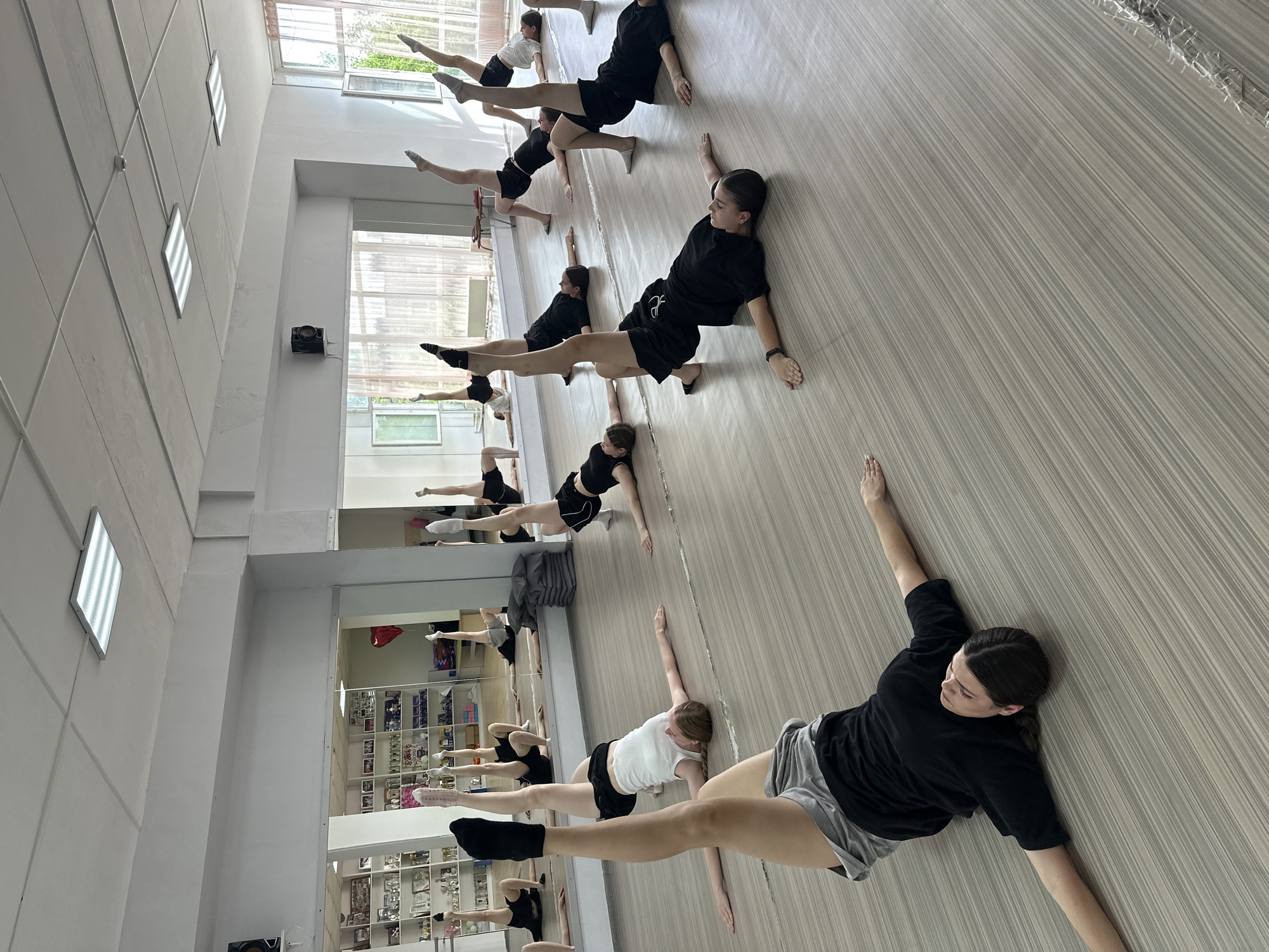 The image depicts a group of dancers in a dance studio. The studio has a light-colored wood floor and large mirrors along one wall, reflecting the dancers and the studio space. The dancers are practicing a stretching or balancing exercise, lying on their sides with one leg extended upwards. They are wearing various dance attire, including black t-shirts and shorts, and socks. The studio appears to be well-lit, with windows visible along the top of the walls. The reflection in the mirrors shows shelves filled with trophies and other items, suggesting the studio has a history and is used for performances or competitions. Based on the available information, it is not possible to confirm the location in Votkinsk, Russia.