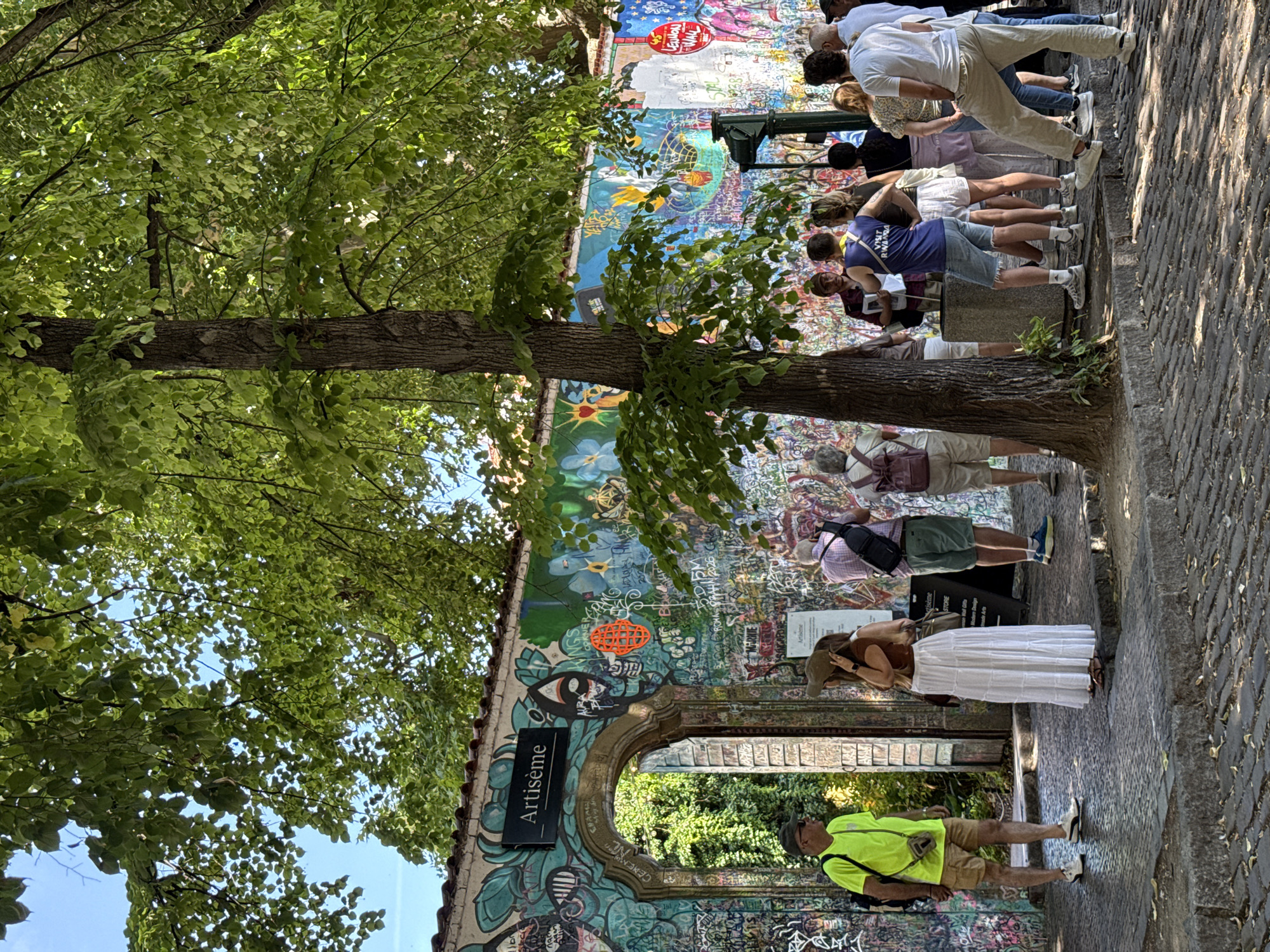 The image depicts a group of tourists gathered in front of a graffiti-covered wall in Prague, Czechia. The wall, prominently featuring the word "Artiseme" in a black sign above an arched entrance, is adorned with colorful murals and an abundance of spray-painted tags and drawings. The arched entrance leads to a lush green area visible behind the wall.
Several people are present, appearing to be tourists engaged in sightseeing. In the foreground, a man in a neon yellow t-shirt and khaki shorts stands to the left, facing away from the camera. To his right, a woman in a white maxi skirt and a brown top stands with her back to the viewer, holding a hat. A group of people are clustered further back, observing the wall and seemingly listening to a guide. One man in a blue tank top with "Visit Rwanda" printed on it is speaking to the group.
The environment is outdoors, with large trees providing shade over the scene. The ground is paved with cobblestones. The overall atmosphere suggests a cultural and artistic attraction within the city. The style of graffiti and the presence of the "Artiseme" sign suggest this location might be related to the John Lennon Wall area, known for its artistic expression and historical significance in Prague.