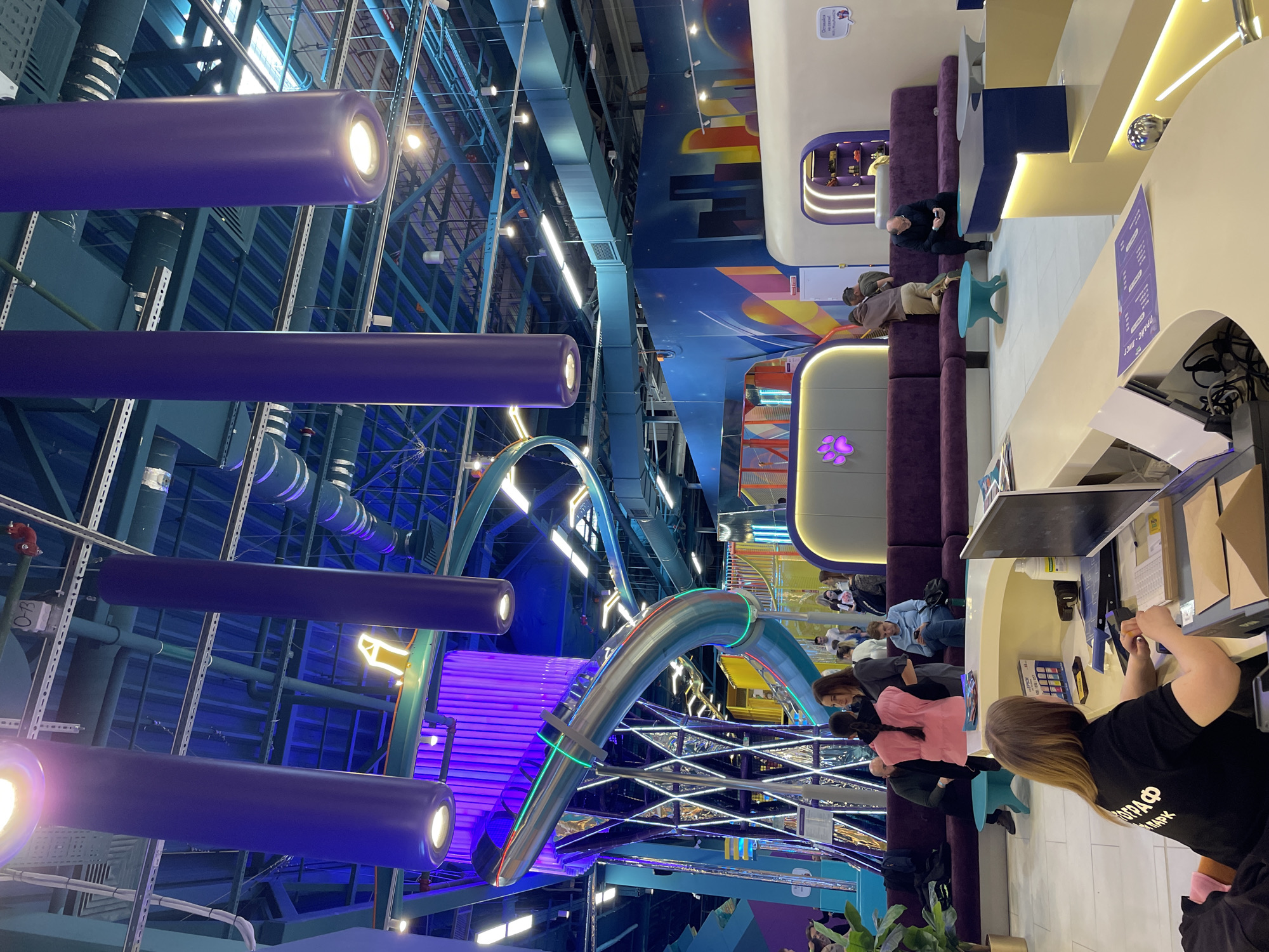 The image captures an interior view of a space in Kazan, Russia. It appears to be a recreational or entertainment area. The environment features a high ceiling with visible structural elements and colorful, abstract decorative elements. There are several suspended light fixtures, cylindrical in shape and purple, with illuminated ends. A prominent architectural feature is a curving metallic structure, accented with green and red stripes.
People are present in the space, seated along a purple upholstered bench against a light-colored wall. One individual is at a counter interacting with what appears to be a computer or payment system. The people are casually dressed, suggesting a relaxed atmosphere.
Other objects in the scene include various abstract decorations on the walls, along with furnishings such as tables and chairs. The overall impression is of a modern, vibrant, and possibly child-oriented interior.