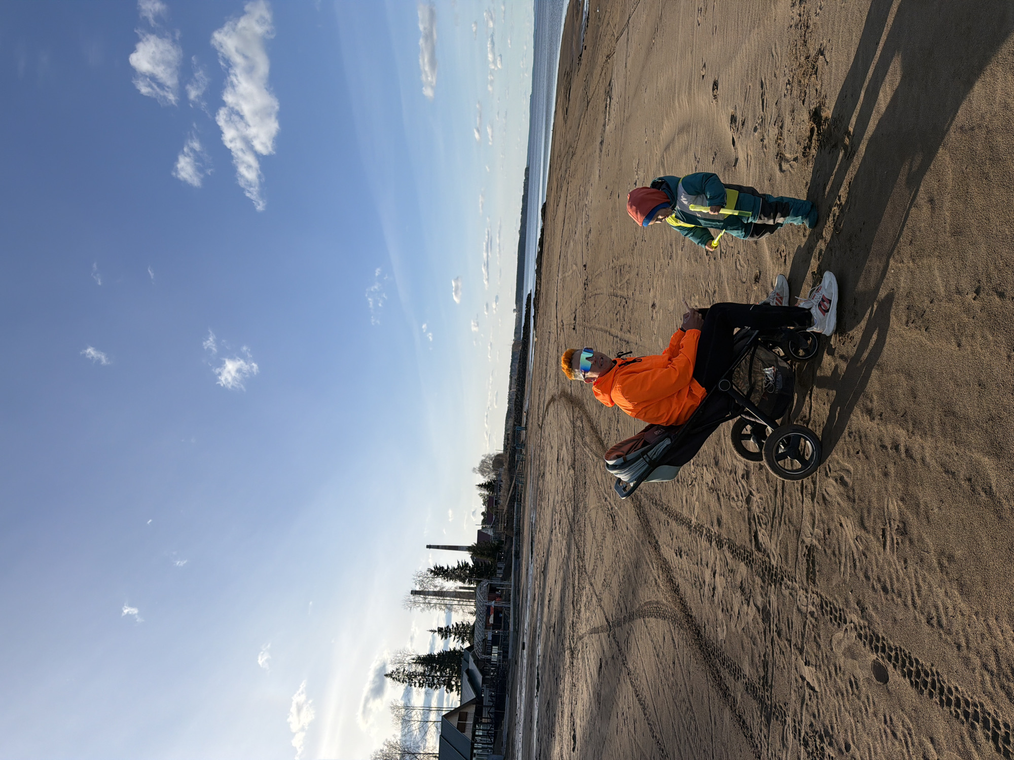 The image shows a person sitting in a stroller on a sandy beach. Another person is standing nearby, facing away from the seated individual. The stroller is casting a long shadow. The beach is sandy with tire tracks visible. In the background, there's a view of buildings and trees. The sky is blue with scattered clouds. Based on the architecture and landscape, this image was most likely taken in the city of Chaikovsky, Russia.