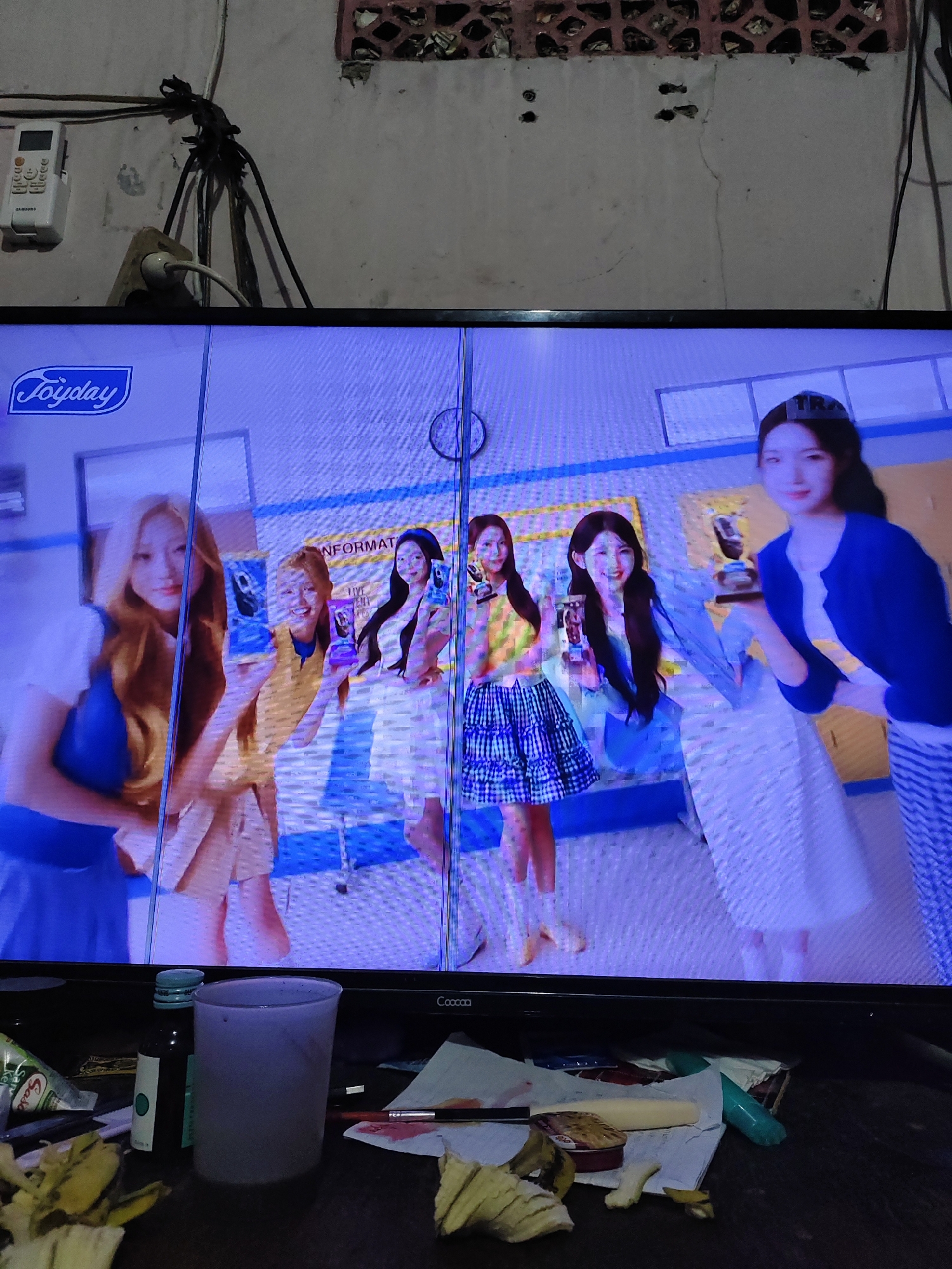 In South Jakarta, Indonesia, an unlit television screen displays a scene of four young women in a bright, indoor setting, possibly a studio or a stylized room. They are dressed in casual, fashionable attire. The woman on the far left wears a blue sleeveless top and a white skirt. The second woman from the left is in a yellow top and a tan skirt. The third woman from the left has long dark hair and wears a blue top and a white skirt. The woman on the far right is in a blue cardigan over a white top and a white skirt with blue stripes. Three of the women are holding up individual items, possibly products or phones, towards the viewer. The woman on the far right holds a trophy-like object. The television screen itself shows a slight vertical distortion line down the middle. In the foreground, on a dark surface, are various objects: a glass, a small dark bottle, a crumpled paper, and some pieces of what appear to be food items, possibly banana peels. The background behind the television shows electrical wires and a portion of a wall with a ventilation grate.
