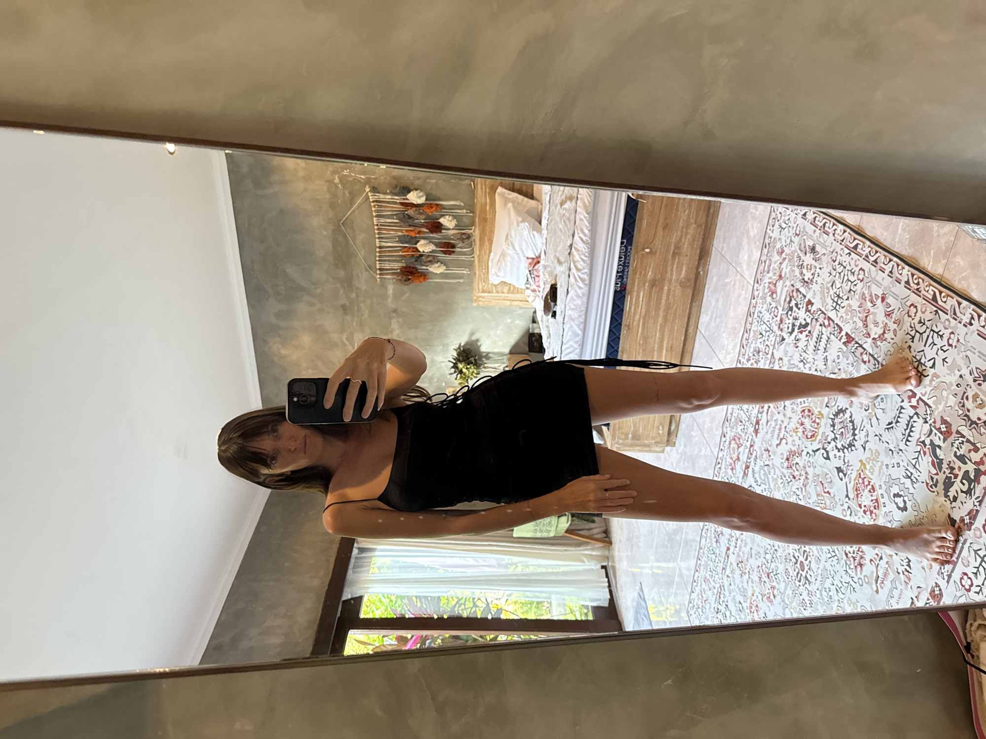 A woman is seen in a mirror selfie, in what appears to be a bedroom. She is wearing a black dress with thin straps. The background includes a bed with white bedding, a wall decoration, a window with greenery visible outside, and a patterned rug on the floor. This scene is purportedly located in North Kuta, Indonesia.
