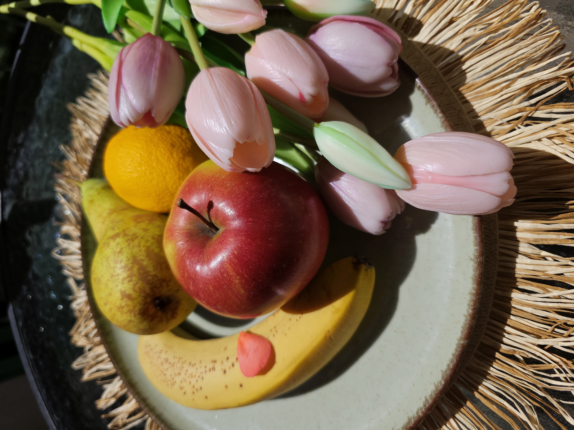 A still life arrangement is presented on a plate with a light greenish hue and a textured rim. The plate is positioned on a surface with straw-like textures extending beyond the plate's edge. The arrangement features a banana with a pink, petal-shaped object adhered to it, a glossy red apple, a bright orange citrus fruit, and a pear with a yellow and brown speckled skin. Above the fruit, there are several tulip buds with pink petals and a single green tulip leaf. The setting lacks discernible location information, rendering the city "Unknown, Unknown."