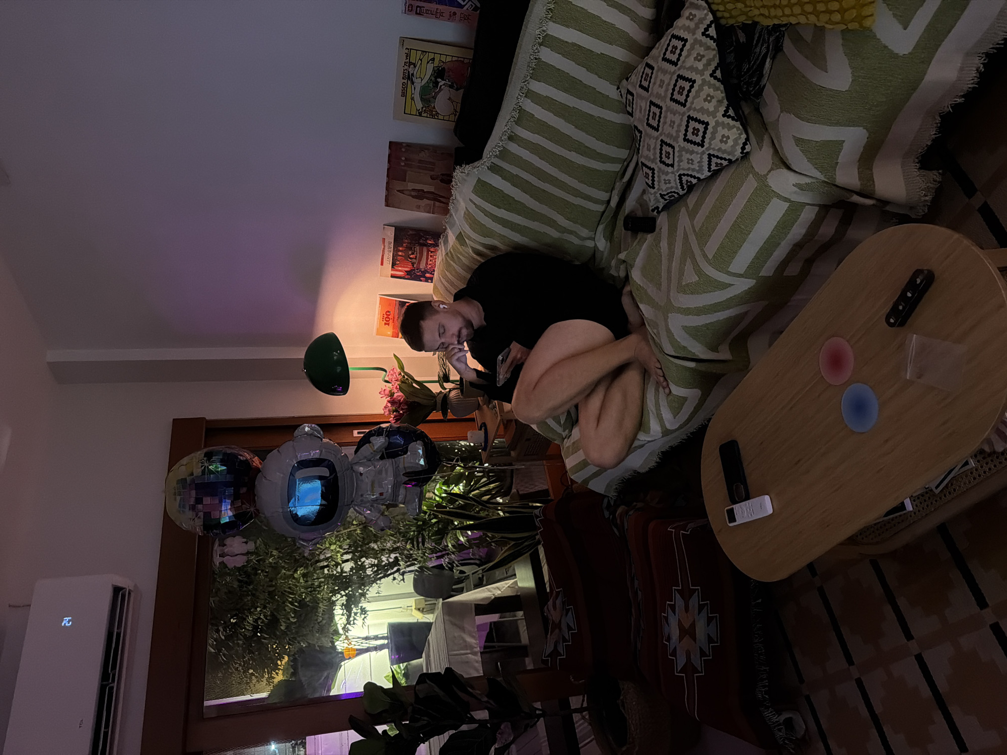 In a dimly lit living room, a man sits on a striped sofa, holding a phone and wearing wireless earbuds. An air conditioner unit displays a temperature of 24 degrees Celsius on the wall above him. In the background, several balloons, including one shaped like an astronaut, float near a window offering a view of greenery and a table set with white cloth. The sofa is covered with a green and white striped throw and decorative pillows. A wooden coffee table in front of the sofa holds a remote control, a phone, and two coasters. The floor is covered with a patterned rug.