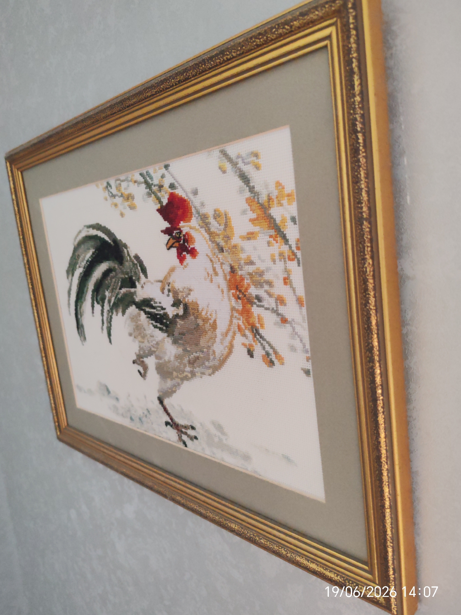The image shows a framed cross-stitch artwork of a rooster. The artwork is predominantly white, with the rooster depicted in various shades of grey, brown, black, and white, featuring a red comb and wattle. The rooster is shown with its tail fanned and is set against a background of branches with yellow and orange blossoms. The frame is ornate and gold-colored, with a muted grey matting surrounding the artwork. The framed artwork is hanging on a textured, light blue wall. The bottom right corner of the image displays a timestamp: "19/06/2026 14:07". The scene appears to be an interior shot, possibly in a home or gallery, with no specific identifiable landmarks of Yablonovsky, Russia visible.