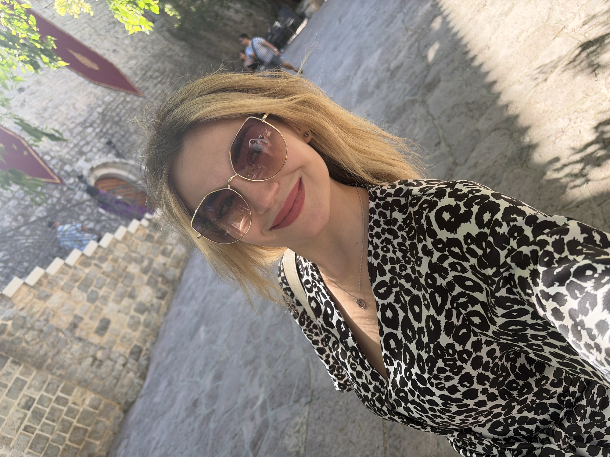 A woman with blonde hair and sunglasses, wearing a black and white animal print dress and a silver necklace, is posing for a selfie. She is standing on stone pavement, with stone steps and a stone building behind her, adorned with maroon banners. Two blurred figures are walking away from the camera in the background. The location appears to be Budva, Montenegro, based on the architectural style.