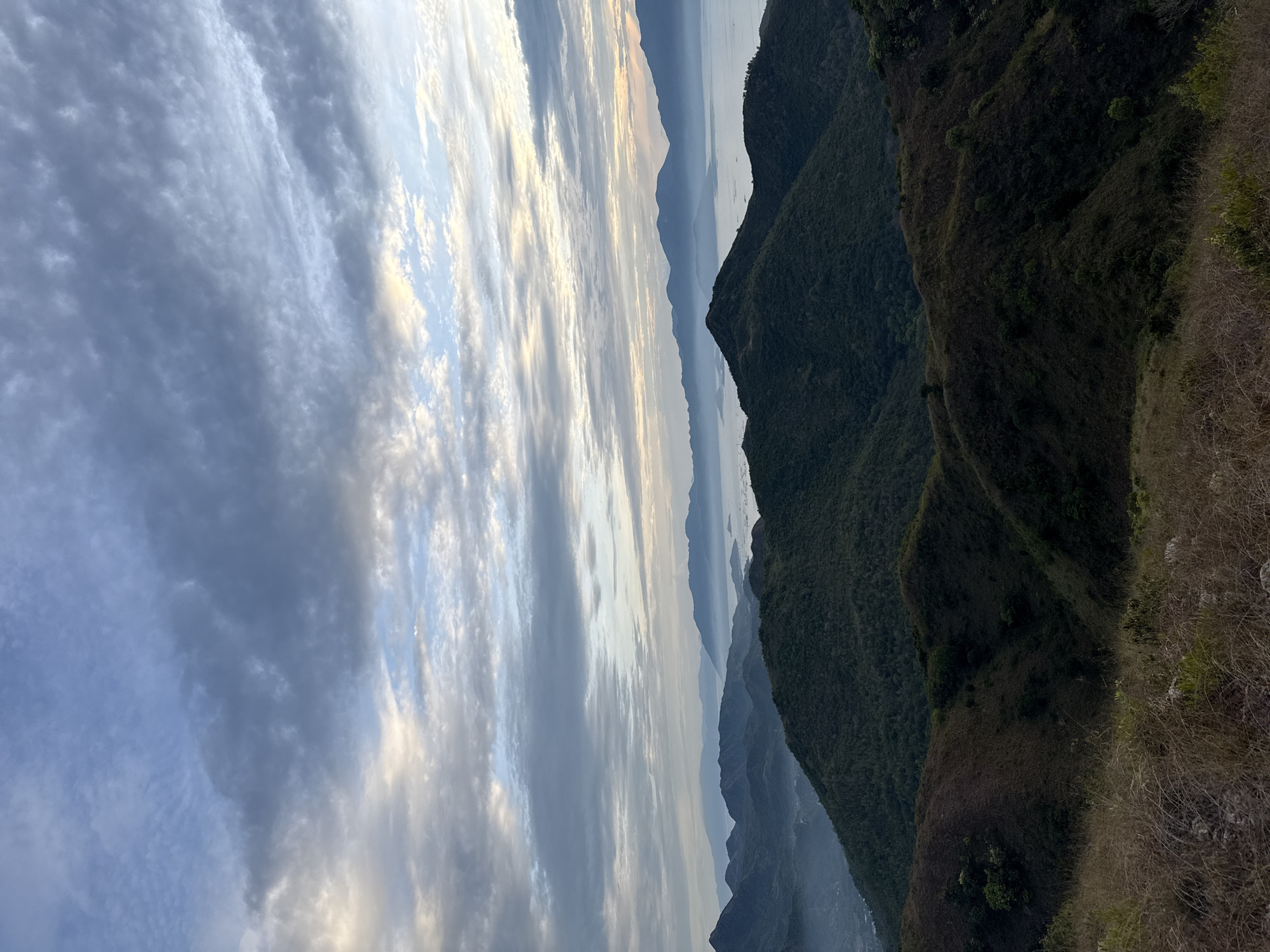 The image depicts a landscape from a high vantage point in Bac Nha Trang Ward, Vietnam. The foreground features a grassy hillside with dry vegetation. In the midground, a series of progressively distant mountains are visible, covered in lush green vegetation that fades to a dark green hue. Further in the distance, a body of water is partially visible between the mountains. The sky fills the left side of the frame with an array of clouds, tinged with varying shades of gray and hints of blue and gold, suggestive of sunrise or sunset.