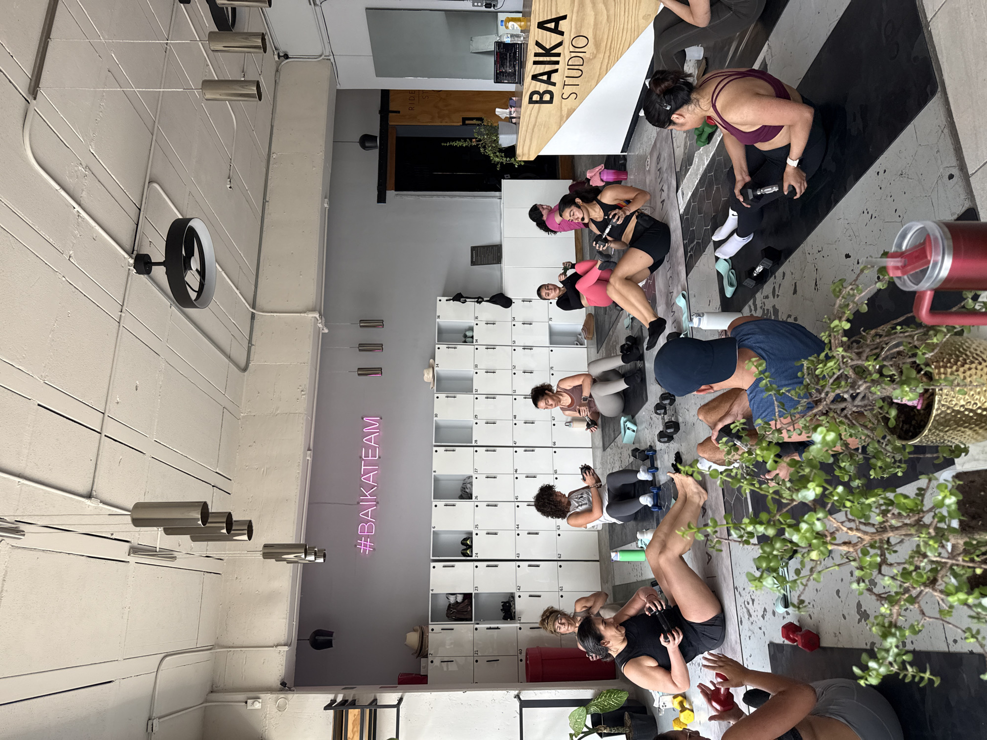 A group of people participates in a workout session at the BAiKA STUDIO, as indicated by the studio's signage. The location appears to be a fitness studio with white lockers along one wall and a neon sign reading "#BAiKATEAM." People are using dumbbells, and exercise mats are positioned on a patterned floor. The setting includes elements of interior design such as overhead lighting fixtures, visible pipes, and potted plants. The location in the city of Puebla, Mexico, cannot be confirmed based on the image alone.