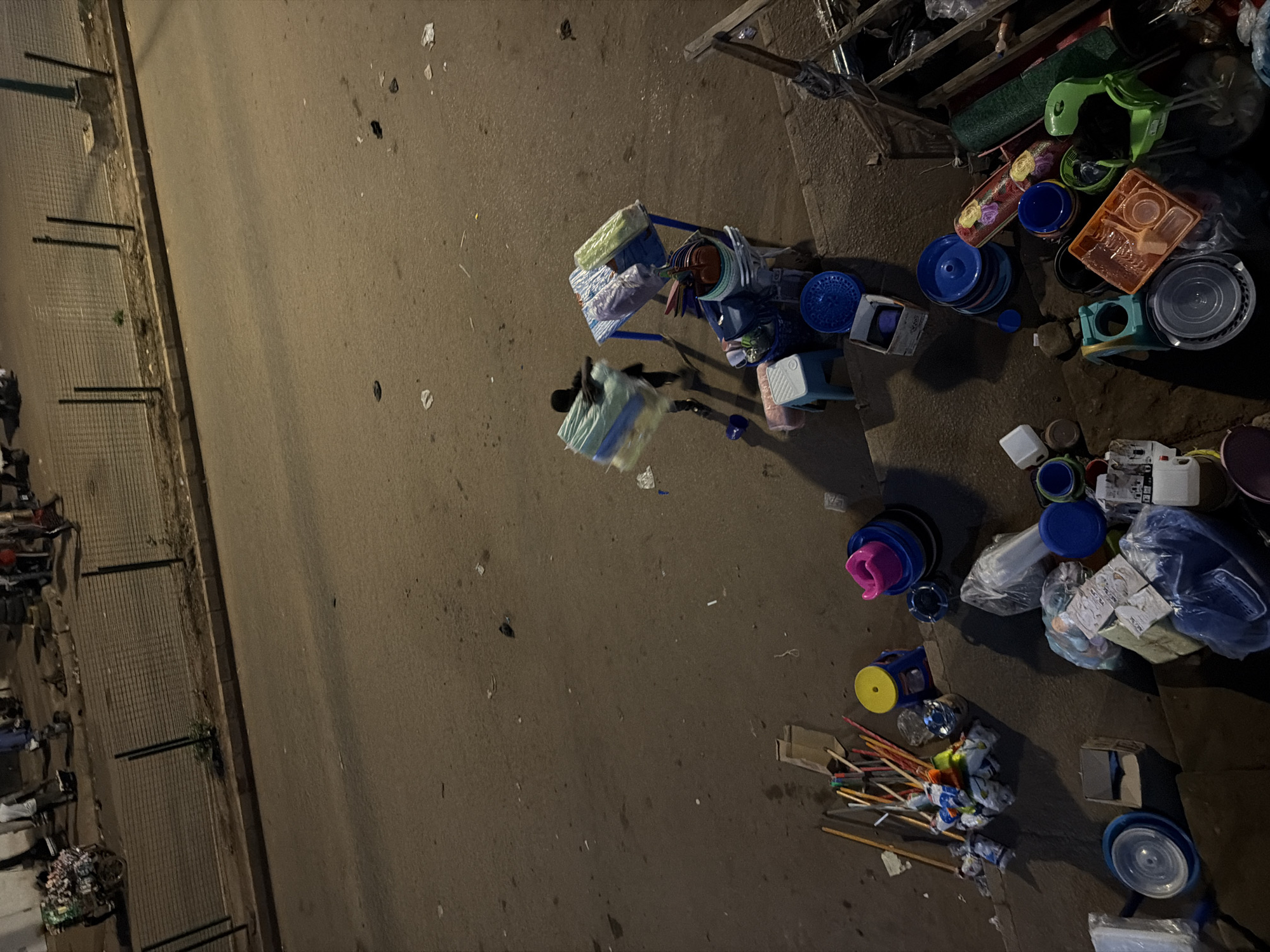 The image depicts a street scene at night in Bukuru, Nigeria. The ground is paved asphalt, with scattered debris like trash and leaves. To the left, a metal fence runs along the edge of the road, with some vegetation growing near its base. The majority of the frame is occupied by a collection of various plastic household items, including buckets, basins, plates, and containers. These items are piled on a blue plastic cart and scattered on the ground. Some of the items are wrapped in plastic. There is a single person visible, dressed in dark clothing and carrying a bundle wrapped in a light-colored plastic bag, walking across the street. Another person is partially visible, hunched over near the cart. The lighting suggests it is nighttime. The overall impression is of a street vendor or someone transporting or selling these goods, with the items accumulated in a somewhat disorganized manner.