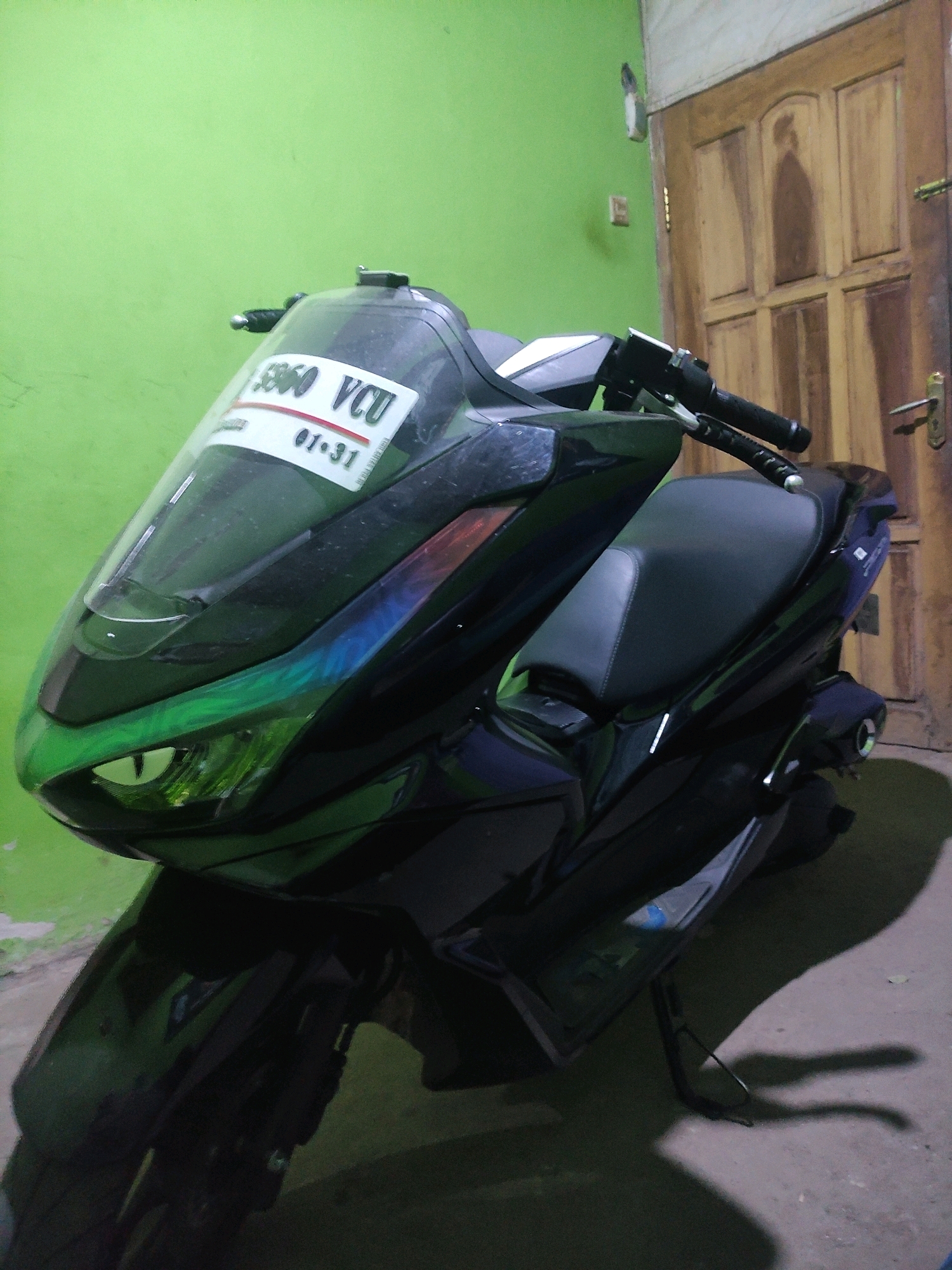 A dark-colored scooter is parked indoors, in front of a wooden door and against a bright green wall. The scooter's license plate reads "3860 VCU" and "01-31". The scooter has a reflective windshield and iridescent accents on its fairing. The floor appears to be concrete, and a shadow of the scooter is cast on it. The scene is dimly lit, likely at night, and the overall impression is of a residential area or garage. There are no people visible in the image.