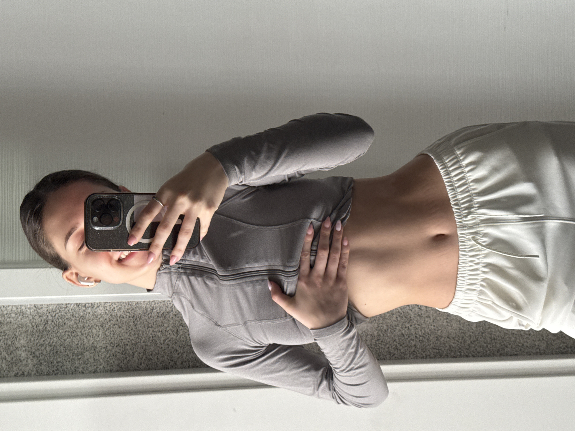 A mirror selfie captures a young person, likely female, smiling broadly. The image shows the subject from the chest down to the upper thighs, taken indoors with a light-colored wall or ceiling in the upper background and a grey carpeted floor in the lower part of the frame.
The person holds a dark-colored smartphone, featuring multiple camera lenses and a visible magnetic attachment on its case, in their left hand, partially obscuring their face. A silver ring adorns the ring finger of the left hand. Both hands display light pink nail polish. A white wireless earbud is visible in the person's right ear.
The subject is dressed in a long-sleeved, grey athletic top with a partial front zipper, which is cropped to reveal a toned midriff and navel. Their right hand rests gently on their abdomen. They are also wearing white, loose-fitting pants with an elasticated, gathered waistband and visible drawstrings.
The smile on the person's face conveys a cheerful and confident mood. No discernible text is visible within the image.