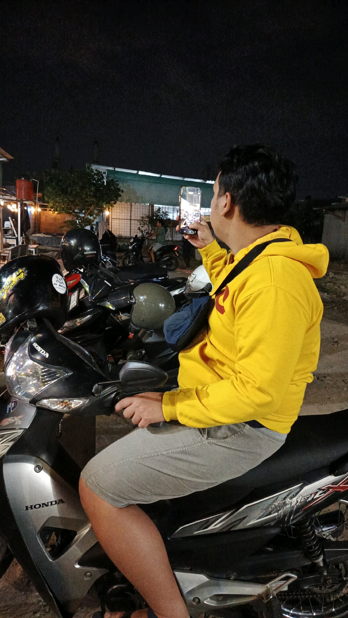 A man wearing a bright yellow hooded sweatshirt and light grey shorts sits on a motorcycle at night in Pabelan, Indonesia. He is holding a smartphone in his right hand, aiming it towards the left, appearing to take a picture or video. The motorcycle is parked alongside several other motorcycles, some of which have helmets on them. The background shows a dimly lit outdoor area with a building structure and trees. The scene suggests a casual gathering or a stop in a public area.