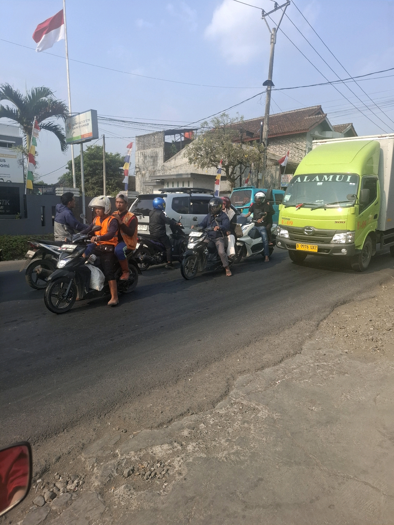 A street scene in Karangtengah, Indonesia, features several motorcycles and a light green truck on a paved road. The road surface shows signs of wear and repair. Two men on a black motorcycle in the foreground are wearing orange vests and helmets. A man in a blue jacket is on another motorcycle to their left. To the right, a man in a black jacket and helmet is on a white motorcycle. Further back, a light green truck with the word "ALAMUI" on its windshield is stopped, with other vehicles and buildings visible in the background under a clear sky. A flag, appearing to be the Indonesian flag, is flying on a flagpole on the left side of the image.