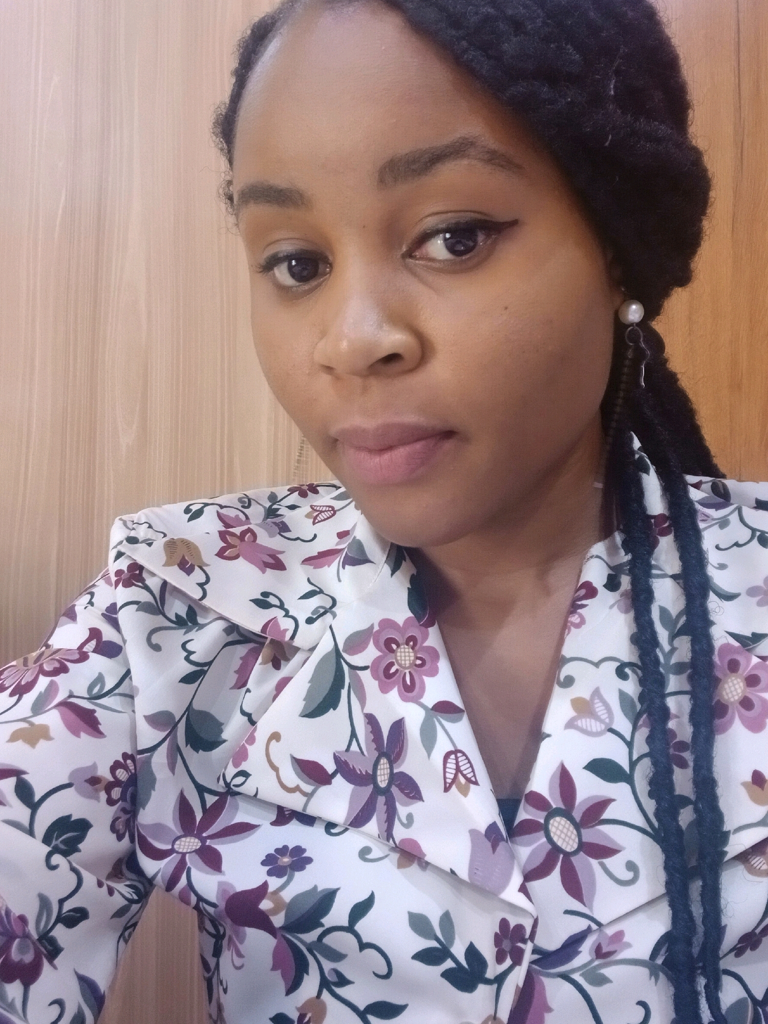 A close-up, medium shot captures a young Black woman with dark braided hair, wearing a white floral print collared blouse. She is positioned in front of a light-colored wooden paneled wall, likely indoors, with the camera at eye level, slightly angled upwards. The woman's face is the primary focus, turned slightly to her left. Her makeup is subtle, featuring winged eyeliner and lip gloss. She wears a pearl earring with a dangling charm in her left ear, and her braided hair is visible over her right shoulder. The floral pattern on her blouse consists of pink, purple, green, and brown flowers and leaves against a white background. The lighting is soft and natural, creating a gentle illumination on her features and the surrounding environment. The overall impression is that of a casual, personal photograph taken in a domestic or office setting.