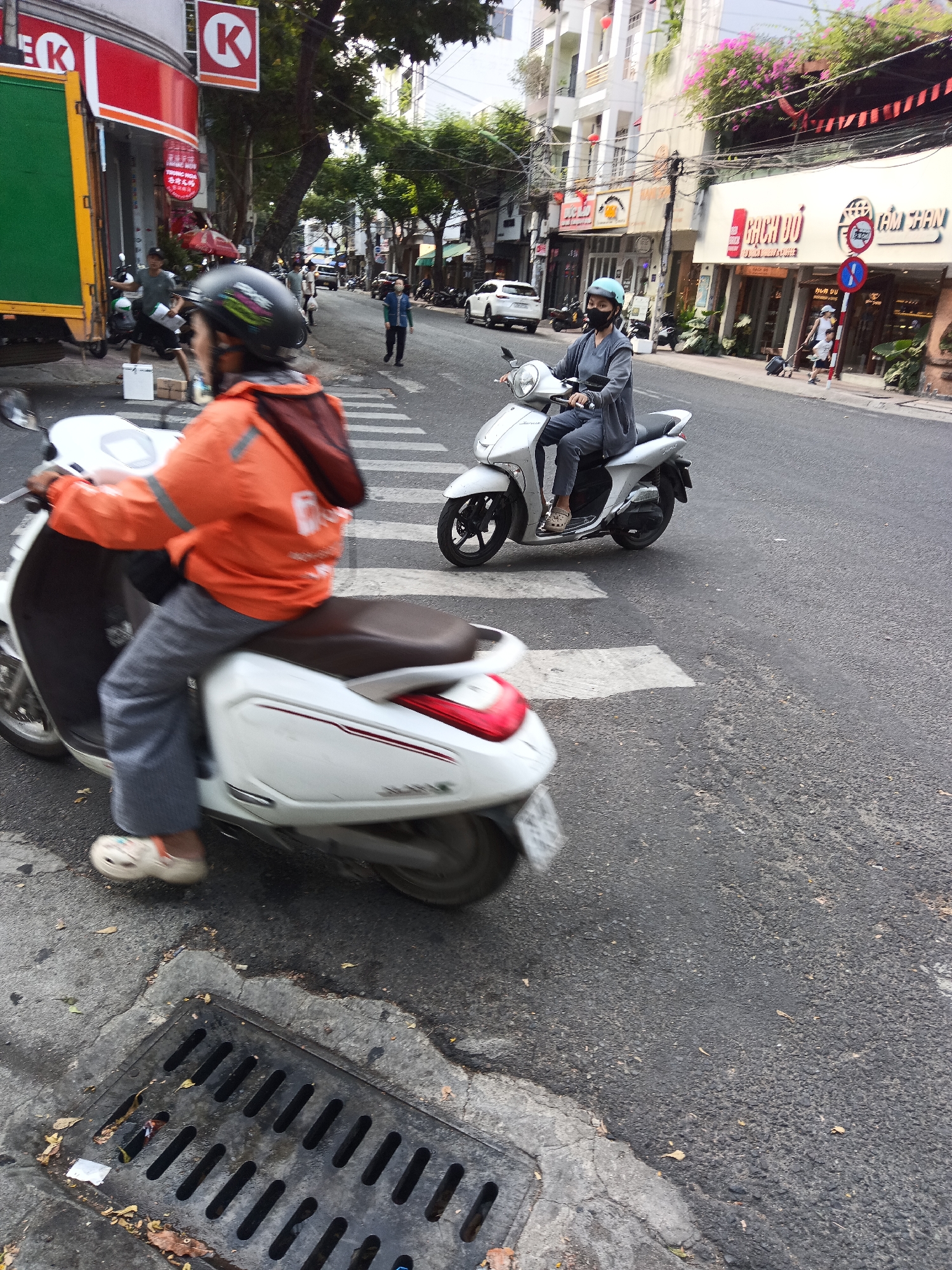 In a city scene in Nha Trang, Vietnam, two individuals are riding scooters on a paved road. The primary focus is on the foreground scooter, ridden by a person wearing an orange jacket and grey pants, with a dark helmet. To the right, a second scooter is occupied by a person wearing a grey jacket, grey pants, and a light blue helmet. The scooters are moving past a pedestrian crossing marked with white stripes. In the background, buildings with multiple stories and various signs are visible. There is a yellow truck with a green container on the left side of the image. Trees line the street, and power lines are strung overhead. A metal grate is visible in the lower left corner of the image. The lighting suggests it is daytime.