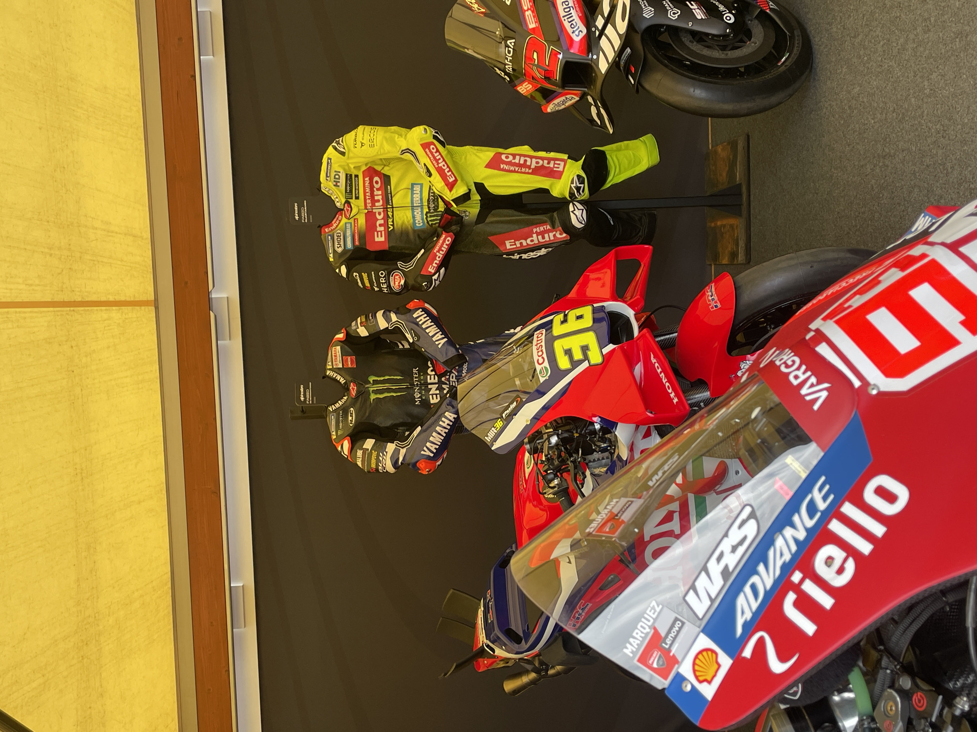 The image shows a display of racing motorcycles and associated racing suits against a black backdrop, likely in an exhibition. The motorcycles are various brands and models, including Yamaha, Honda, and Ducati, indicated by visible logos and team names. Racing suits with sponsor logos (Monster Energy, Enduro, Pertamina, and others) are displayed on mannequins, and are in the style of Valentino Rossi (indicated by "VR46"). The image also features a motorcycle fairing with the number "36" and the names "Marquez" and "Riello", suggesting a connection to Marc Marquez. The general ambiance suggests an event or museum display of motorsports memorabilia, possibly in Barcelona, Spain, as "BARCELONA" is visible on one of the labels.