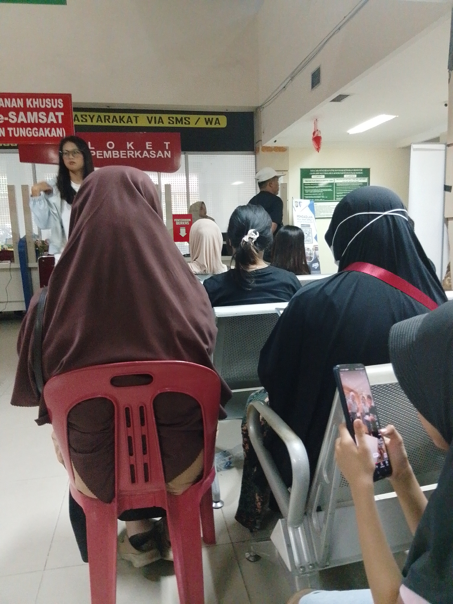 The image depicts an indoor waiting area, likely a government office or service center, in Batam, Indonesia. In the foreground, a person is seated with their back to the camera, wearing a maroon hijab and black pants. They are sitting on a red plastic chair. To the right, another person, also with their back to the camera, is wearing a black hijab and clothing with a red strap across their chest. This individual is holding a smartphone and appears to be looking at it. Further back, several other individuals are visible, mostly seated and facing away from the camera. Some are wearing hijabs in various colors, while one person in a white cap is standing and facing to the left. The environment includes rows of chairs, a counter area with signs, and fluorescent lighting. One prominent sign in red and yellow lettering reads "ANAN KHUSUS E-SAMSAT MASYARAKAT VIA SMS / WA" and "LOKET PEMBERKASAN." Another sign to the left has the text "POLDA KEPRI." The overall atmosphere suggests a public service facility.