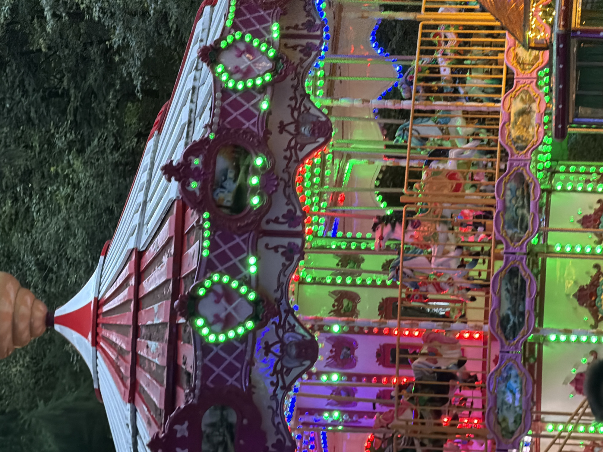 The image shows a close-up, vertically oriented view of a carousel at an outdoor amusement park, possibly at dusk. The carousel features a red and white striped conical roof on the left, with an orange, balloon-like structure attached to its apex. The main body of the carousel is adorned with decorative purple and gold trim, illuminated by numerous bright green, blue, and red lights. Intricate patterns and oval-shaped panels, some containing what appear to be painted scenes, are visible on the carousel's structure. Behind the carousel, a dark, leafy background suggests trees, hinting at a park setting.
The carousel itself is a complex structure with multiple levels and a golden metal framework. Within this framework, carousel horses and other ride elements are partially visible, suggesting that the ride is either stationary or in motion. The vibrant, flashing lights create a festive and somewhat magical atmosphere, common for amusement park rides, especially when illuminated after dark.
There are no visible people in the foreground, though faint figures might be glimpsed within the painted panels or amongst the carousel's internal structure. The time of day appears to be evening or night, given the artificial lighting being the primary illumination source, contrasting with the dark background. The weather cannot be determined from the image. No text is visible. The location is identified as Perm, Russia.