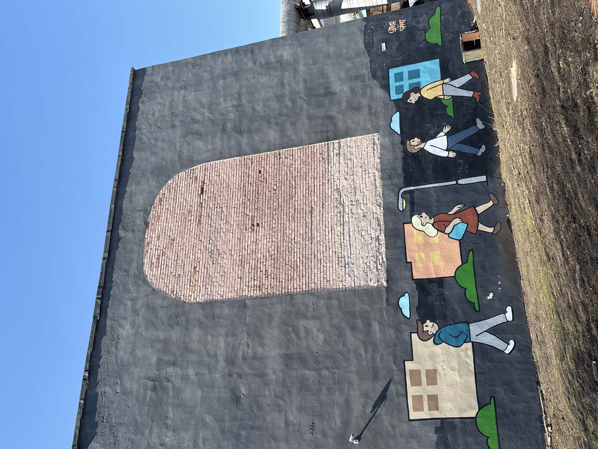 The image shows a portion of a building wall in Izhevsk, Russia, depicting a painted mural. The wall is painted a dark gray color except for a brick-filled arch near the left center. To the right of the arch is a mural depicting cartoon-like figures appearing to walk in an urban setting. The mural includes a woman with blonde hair and a red dress, a man in a white shirt and blue pants, and a person in a yellow shirt and gray pants. Other elements of the mural include houses, trees, and a lamppost. To the right of the mural is overgrown vegetation. The sky above the building is clear and blue. At the very top of the image, there are industrial pipes.