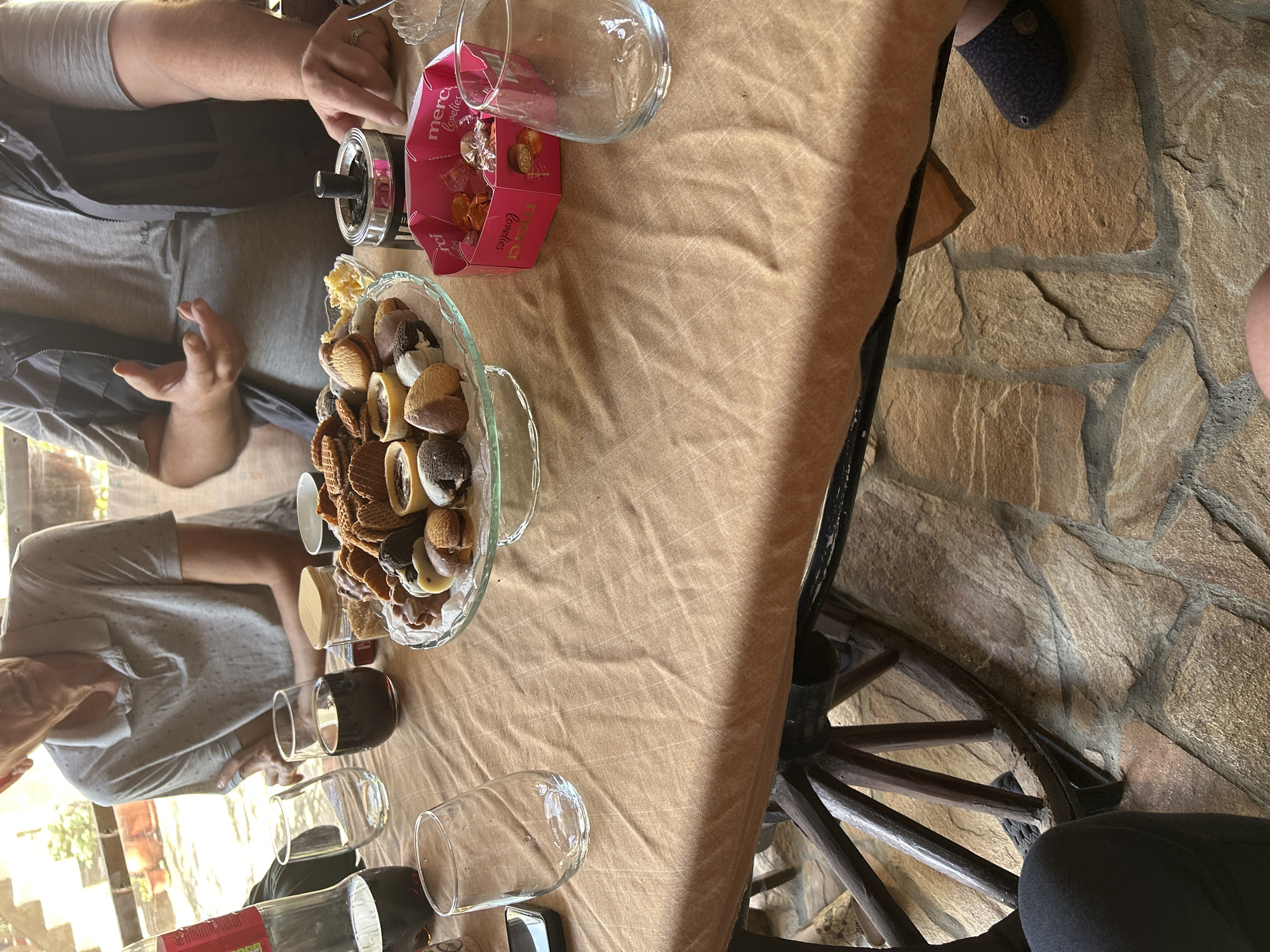 The image depicts a gathering around a table in Bukovlak, Bulgaria. The table is covered with a tan cloth, and it holds a glass bowl filled with assorted cookies and chocolates, a pink box of "Merci Lovelies" chocolates, several wine glasses, a bottle of wine, and an ashtray. Three people are visible partially in the shot. One is wearing a gray shirt and a dark vest, another wears a gray shirt, and a third shows a foot with a slipper. The table appears to be attached to a decorative wooden wheel. The floor is made of stone tiles. The scene suggests a casual, possibly outdoor, setting with people enjoying refreshments.