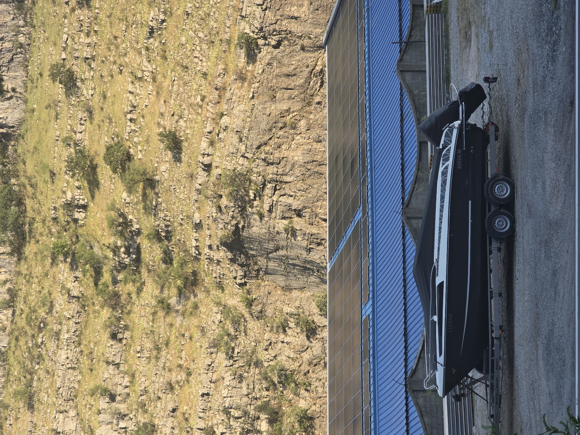 In Podgorica, Montenegro, a black and white motorboat rests on a trailer in an outdoor storage area. The boat is partially covered with a black tarp. Behind the boat, a building with a blue corrugated metal roof is visible, topped with rows of solar panels. Beyond the building, a steep, rocky hillside covered in sparse vegetation rises towards the sky. The ground in the foreground is gravel.